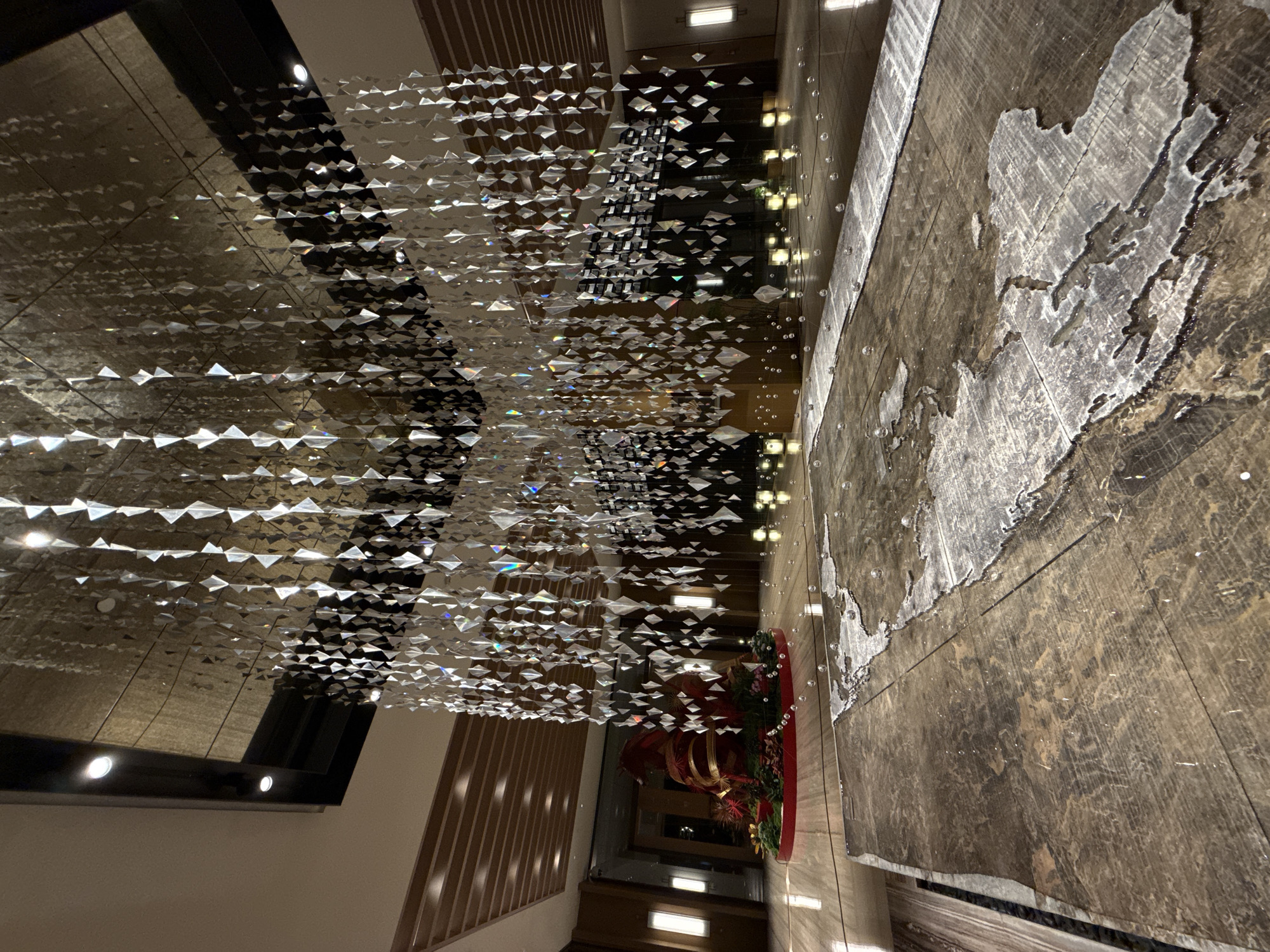 The image shows an interior view of a spacious, modern lobby, possibly a hotel or commercial building in Futian District, China. A large, abstract installation hangs from the ceiling, composed of numerous reflective, diamond-shaped pieces suspended by thin wires, creating a shimmering, cascading effect. The floor is made of polished dark stone with a marbled pattern. In the foreground, a large, irregular metallic or stone inlay resembling a map or abstract topographical feature is set into the floor. Scattered across the floor are small, spherical, reflective objects, possibly decorative or part of the installation. In the background, a vibrant red and gold decorative display featuring foliage and a large, bulbous object is situated on a raised circular platform. The walls are paneled with wood-like material, and vertical strip lighting is visible. The overall impression is one of a sophisticated and artistic interior design. There are no people visible in the image.