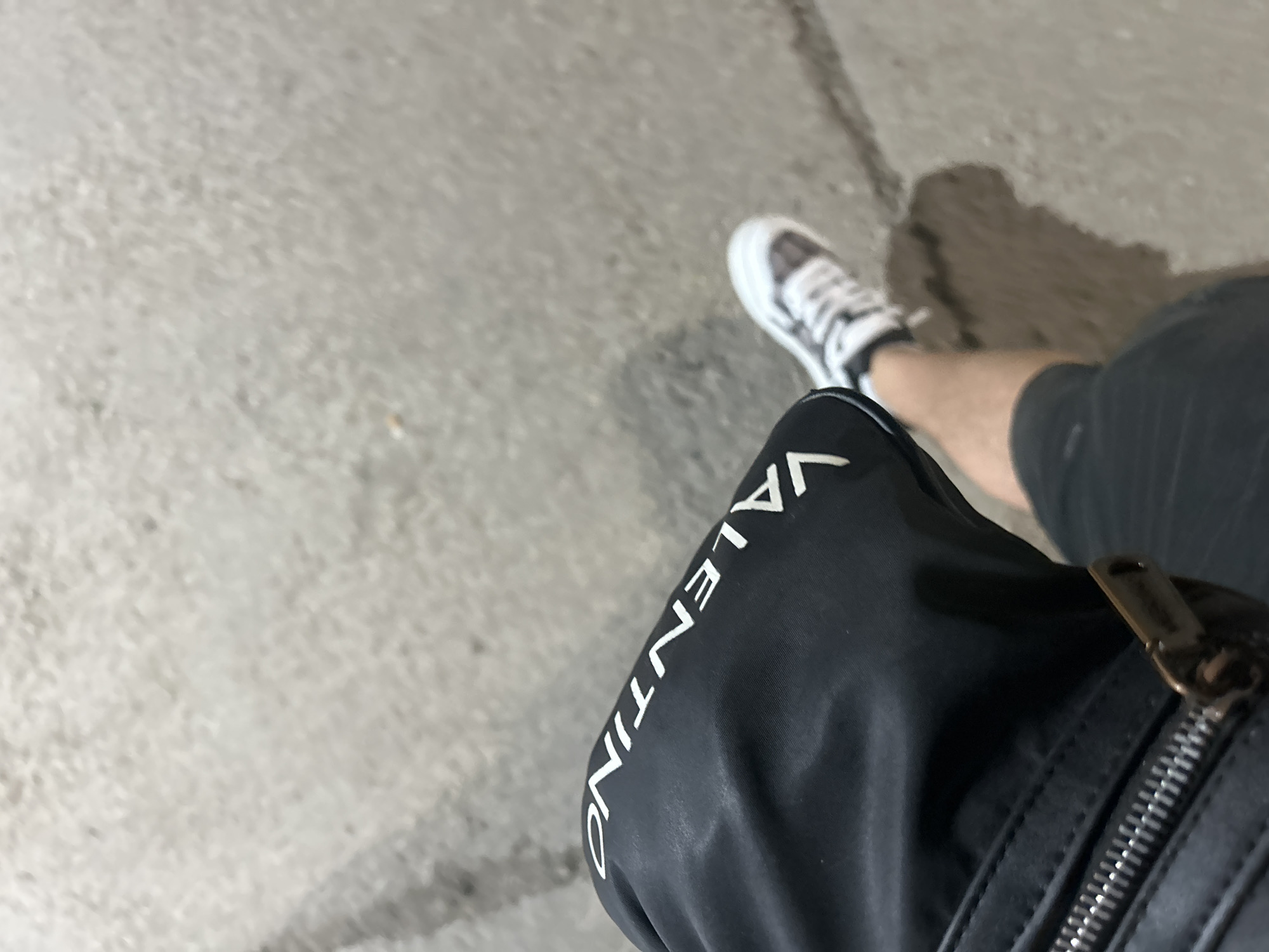 A low-angle, medium shot captures a person's legs and feet on a textured, grey pavement. The person is wearing black shorts, a black bag with white lettering that reads "VALENTINO", and white and grey sneakers. The bag is worn on the person's left hip, with a zipper visible. The person is standing and appears to be walking. The lighting suggests an outdoor setting, possibly during the day.