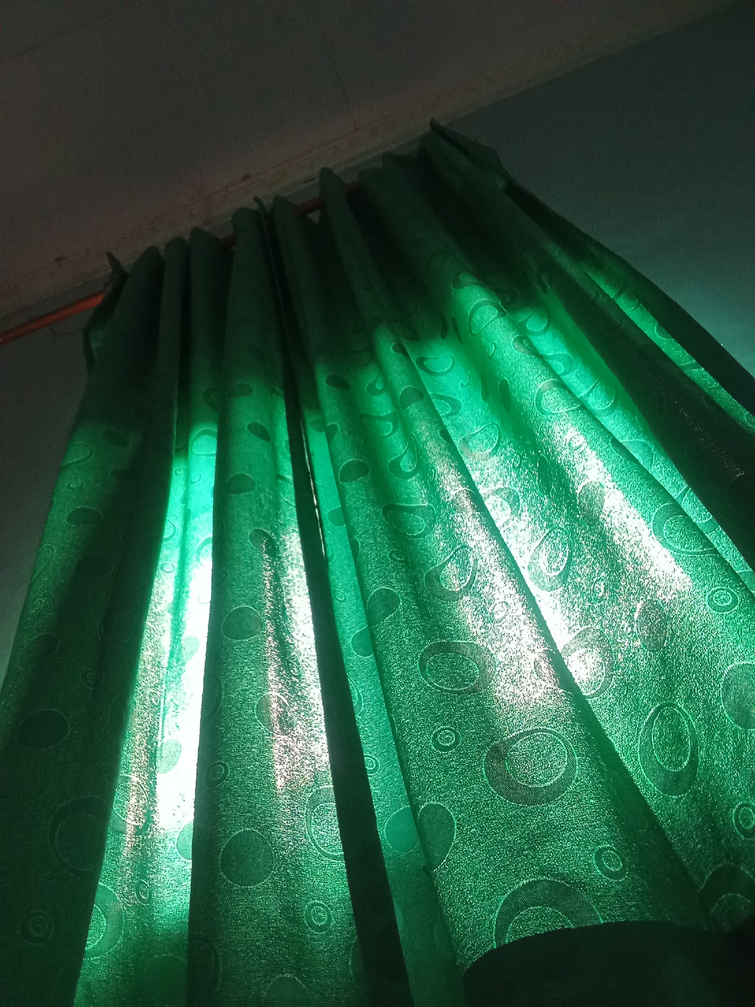 The image shows a close-up view of green patterned curtains hanging indoors. Sunlight is filtering through the fabric, creating bright, vertical streaks of light and highlighting the circular patterns on the material. The top edge of the image shows the ceiling and the curtain rod. The location is identified as Cempaka, Indonesia, but no specific urban features are visible in the image to confirm this or provide further environmental context within the city. The scene is static and focuses solely on the texture and light play on the curtains.