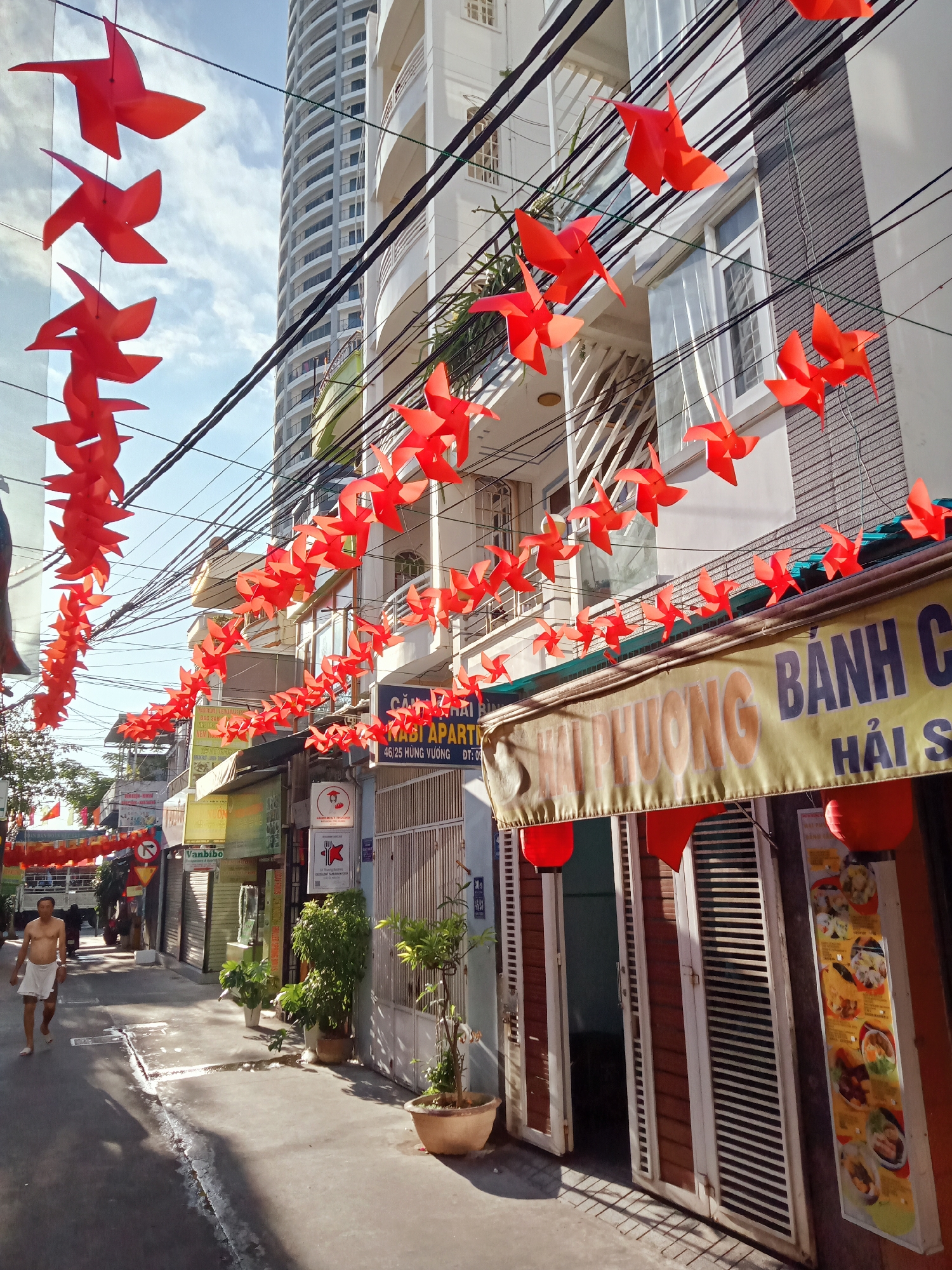 A street scene in Nha Trang Ward, Vietnam, shows a narrow street decorated with red paper pinwheels strung across the electrical wires overhead. The street is lined with buildings of varying heights and architectural styles. A man, shirtless and wearing white shorts, walks along the left side of the street. Some shops are open, indicated by open doors, while others are closed with metal shutters. The "Hai Phuong Banh Canh" sign in yellow and blue is prominently displayed above an open storefront, along with hanging red lanterns. Potted plants are arranged along the sidewalks, adding touches of greenery to the urban scene.