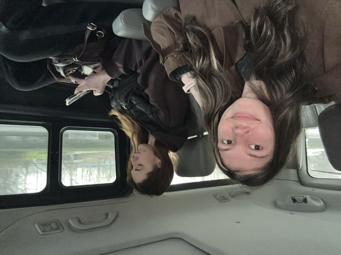 Two young adult females are depicted as passengers in the rear compartment of a vehicle in Huangdao District, China. The vehicle's interior features gray upholstered seating, headrests, and ceiling panels, contrasted with dark window frames.
The female positioned in the foreground, seated on the left, has long, wavy dark brown hair with lighter highlights. She wears a brown jacket with a suede-like texture over a dark green garment. A small stud piercing is visible on her left nostril, and a light-colored smartwatch is present on her left wrist. She is looking slightly to her right, displaying a subtle smile, with her right arm resting on the adjacent seat's armrest.
The second female, seated further back on the right, has long, straight reddish-brown hair. She is dressed in a dark brown long-sleeved top, a black quilted vest, and black pants. Her attention is focused downward on a silver-colored smartphone, which she holds with both hands. A dark brown handbag with gold-colored hardware rests on her lap.
Through the vehicle's windows, an overcast outdoor environment is visible, characterized by trees with sparse foliage, green ground cover, and distant light-colored structures, indicative of an urban or suburban setting. No direct interaction is observed between the two individuals.