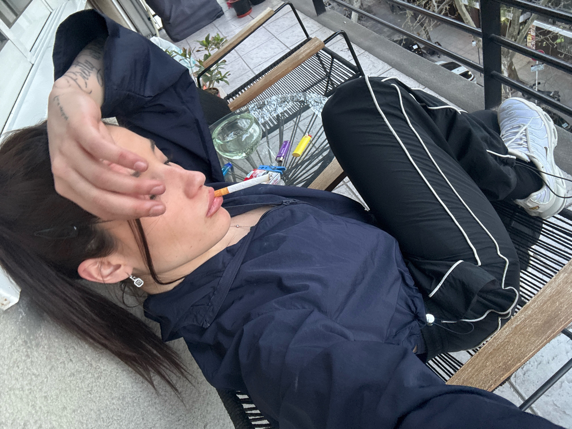 The image shows a person smoking a cigarette on a balcony in Buenos Aires, Argentina. The person is lying on a chair with their hand over their face. There is a glass table in front of them with lighters and an ashtray. The balcony overlooks a street with cars and trees. The person is wearing a navy blue jacket and black pants with white stripes. They have a tattoo on their arm and are wearing a silver earring. The lighting suggests it is late afternoon or early evening.