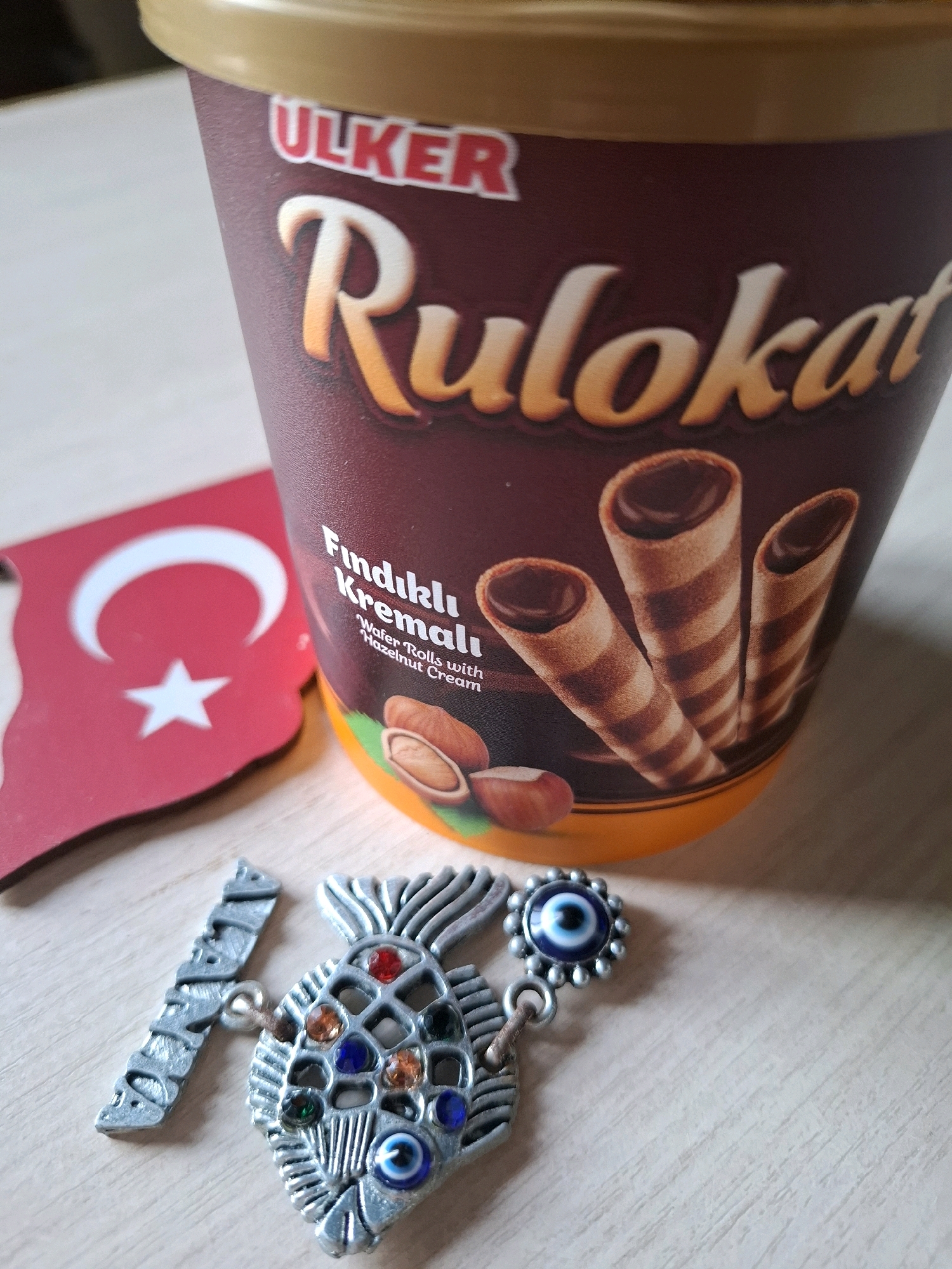 An overhead shot shows a maroon and gold cylindrical container of Ulker Rulokat hazelnut cream wafer rolls, positioned on a light brown wooden surface. To the left of the container is a cutout of the Turkish flag. In the foreground, two small metallic charms are visible: one shaped like a fish adorned with colorful jewels and an evil eye amulet, and another metallic charm that appears to be a letter or symbol. The scene is brightly lit, casting soft shadows on the surface. The image does not contain any identifiable elements that would suggest a specific location in Tyumen, Russia.