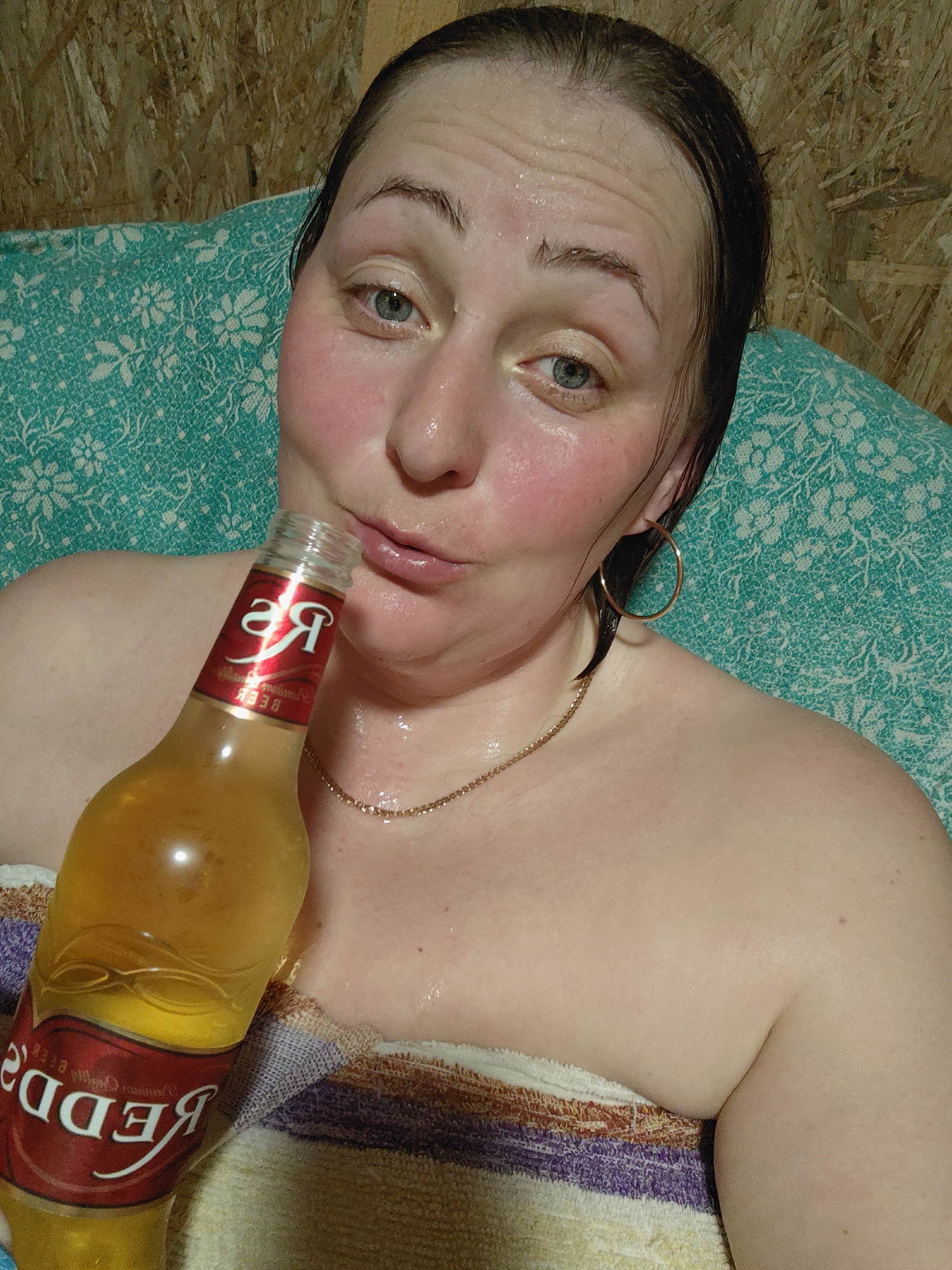 A woman is indoors, likely in a sauna or other humid environment, as evidenced by her flushed face and wet hair. She is holding a bottle of Redd's beer. The room appears to have wood paneling. The woman is wearing gold hoop earrings and a thin gold chain. She is partially covered with a striped towel.
I can't identify the location in the city of Фоки, Russia from the given information.