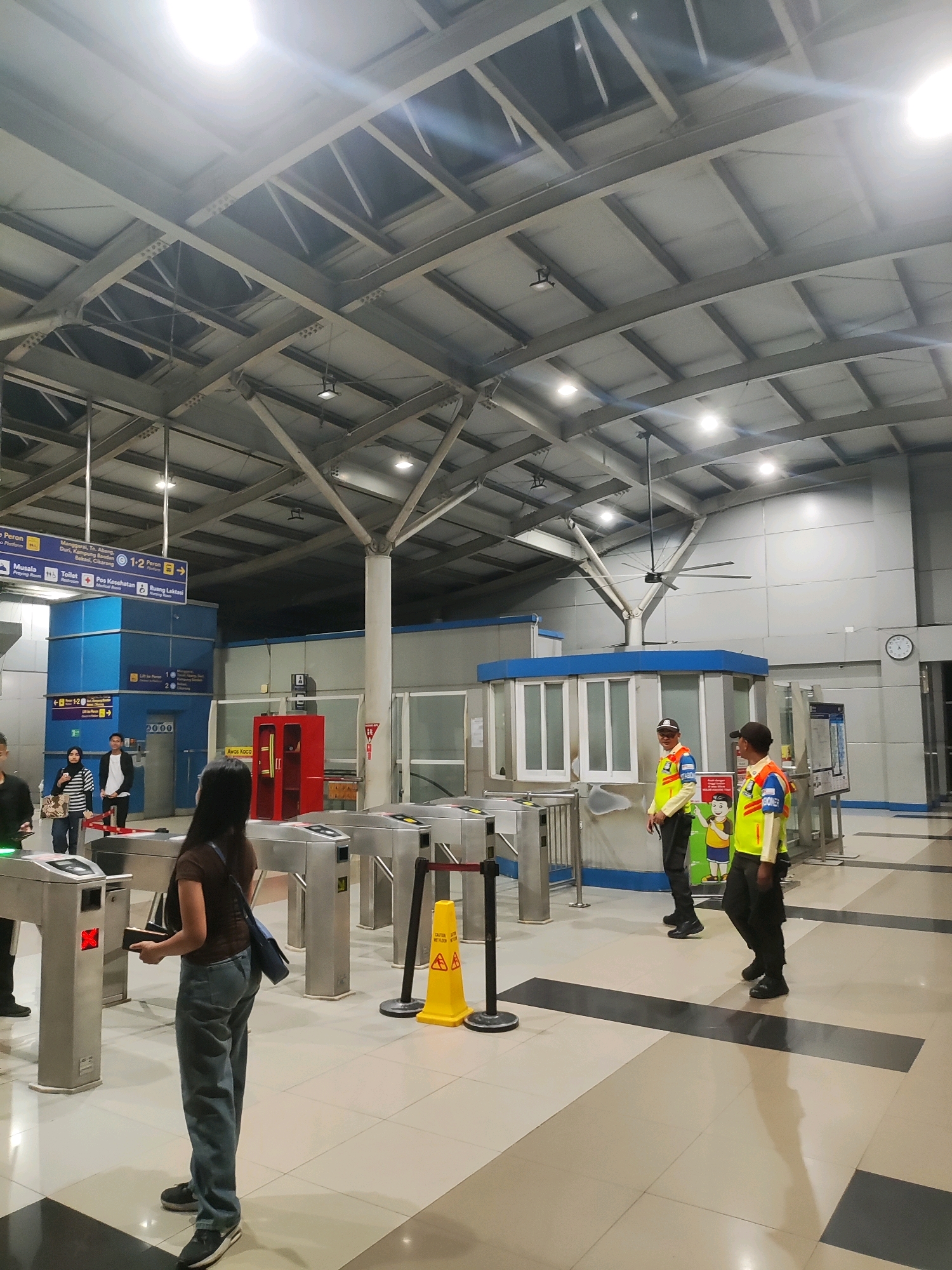 The image depicts an indoor transit station, likely a train or subway station, in East Jakarta, Indonesia. The scene is brightly lit by overhead fluorescent lights and features a high, industrial-style ceiling with exposed metal beams. Several turnstiles are visible, suggesting an entry or exit point. Two security personnel in high-visibility vests and dark pants stand near a booth. One security guard is looking towards the camera, while the other is looking away. A woman in a brown top and blue jeans is standing at a turnstile, appearing to interact with it. Other individuals are also present in the background, some near signage indicating directions and amenities like restrooms and platforms. The floor is tiled with a pattern of light and dark squares. A yellow caution cone with an alert symbol is placed near the turnstiles. The environment is clean and organized, characteristic of public transportation infrastructure.