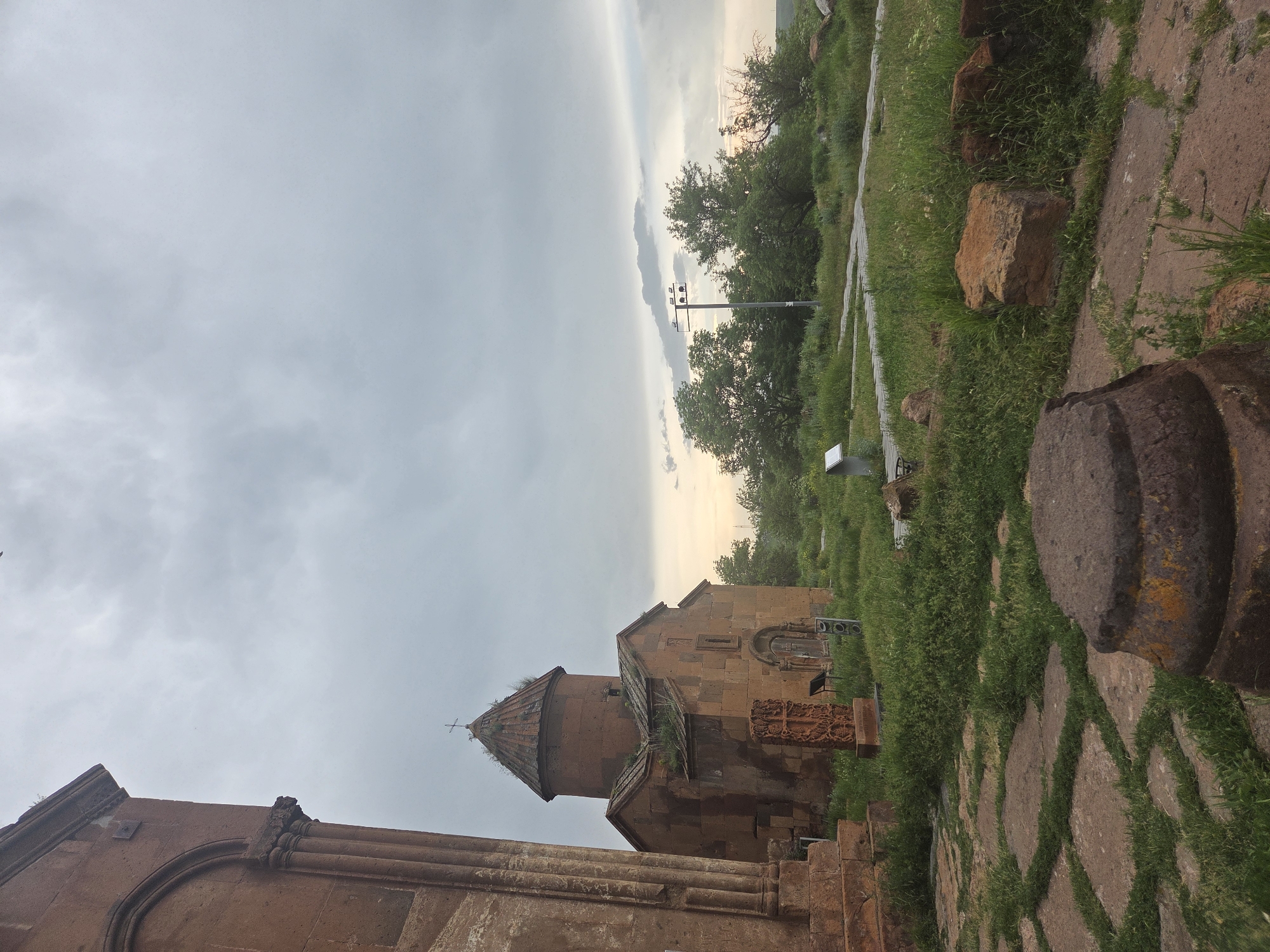 The image depicts a serene, overcast scene at a historical site in Vahramaberd, Armenia. In the foreground, a weathered stone column base, possibly part of an ancient structure, rests on a path paved with large, irregular stone slabs interspersed with grass. Beyond this, a small, old stone church with a conical roof and a cross at its apex stands prominently, its reddish-brown stone facade showing signs of age and vegetation growth. In front of the church, a carved stone monument, resembling a khachkar, is visible. The surrounding area is covered in lush green grass and scattered rocks, with trees and bushes forming a backdrop. A lamppost with multiple lights stands further in the distance, near a path. The sky is a mix of gray clouds and soft, diffused light, suggesting either early morning or late evening. There are no people or animals present in the image.
