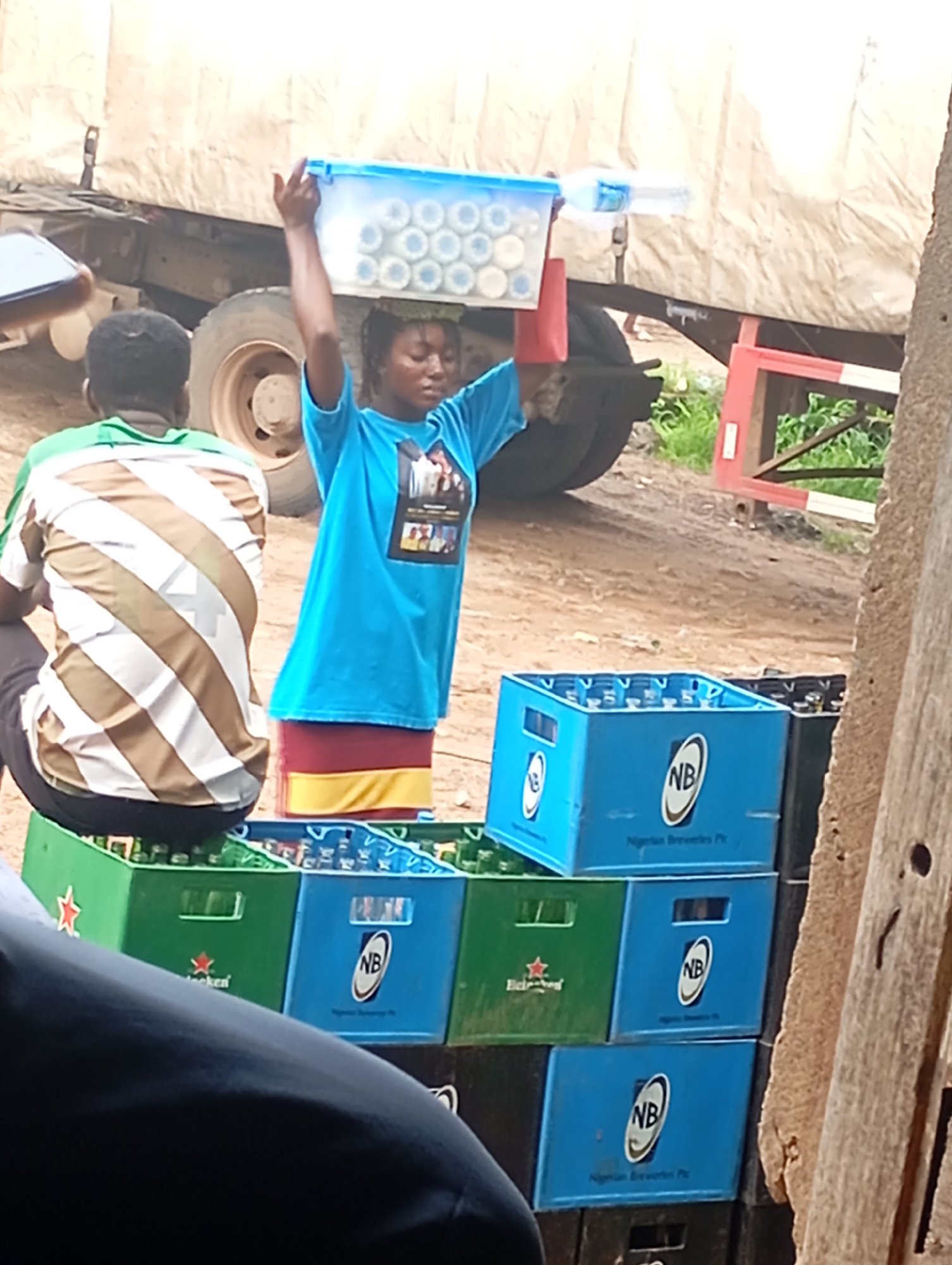 The image depicts a street scene in Shendam, Nigeria, possibly near a market or transport hub. In the foreground, a woman balances a blue plastic container filled with bottled drinks on her head. She is wearing a bright blue t-shirt with a picture on it and a red and yellow skirt. Behind her, a person wearing a brown and white striped shirt is seated. In the background, a large truck is parked. Several crates of bottled drinks, some green and some blue with "NB" logos, are stacked on the ground. The environment appears to be dusty and unpaved, suggesting an outdoor, possibly informal, commercial setting.