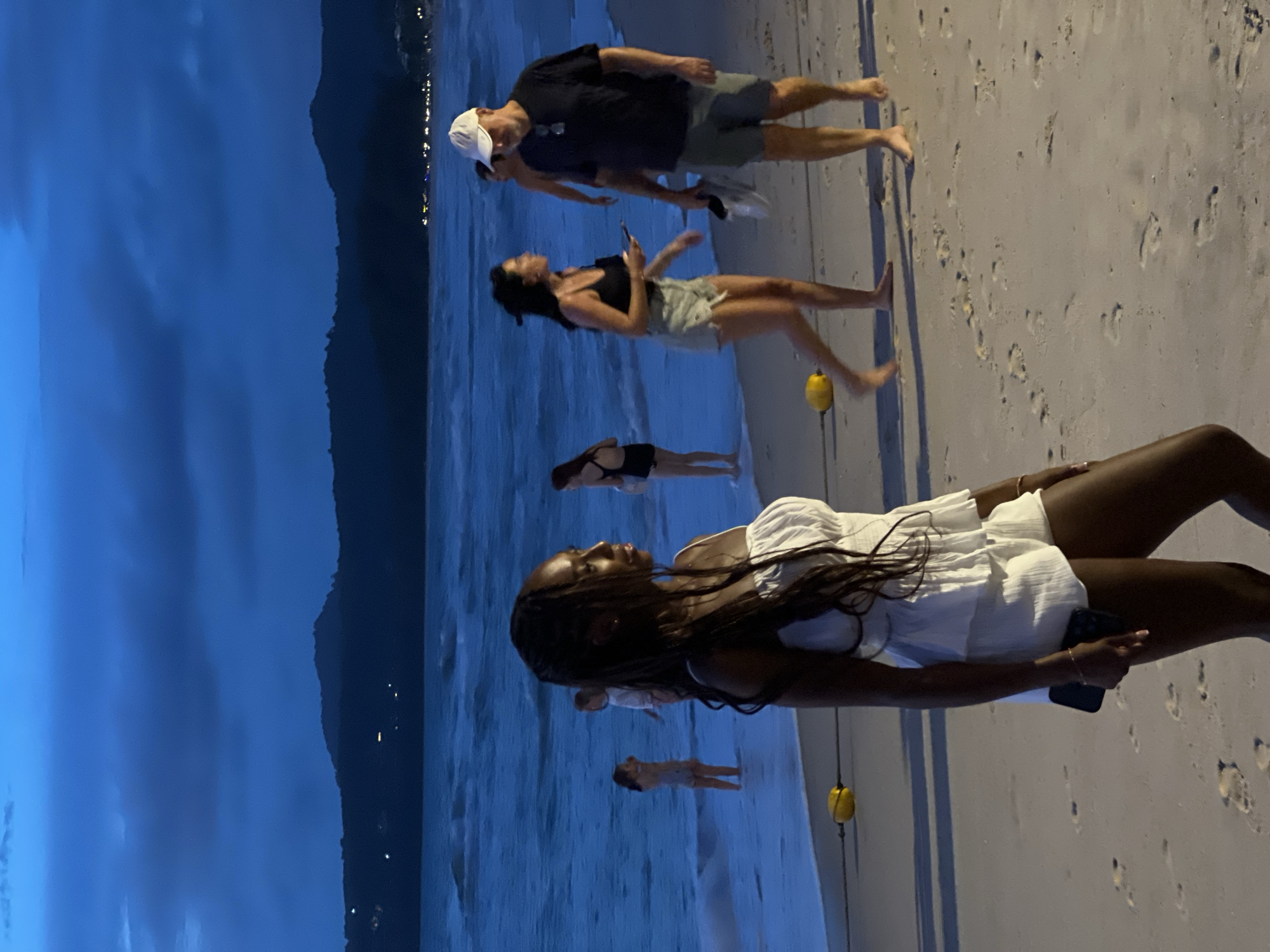 In the city of Unknown, Unknown, at dusk, a group of people are on a sandy beach next to a calm, blue ocean. In the foreground, a Black woman with long, dark hair wears a white outfit and holds a phone in her right hand. Behind her, to the left, two young girls in swimwear stand in the shallow water. Further back, a woman in a black tank top and denim shorts talks to a man wearing a dark t-shirt and a white baseball cap. The man is holding a pair of shoes. The ocean stretches out to the horizon, where distant hills are silhouetted against a dark blue sky with visible clouds. Yellow buoys are placed along the shoreline, and footprints are scattered across the sand.