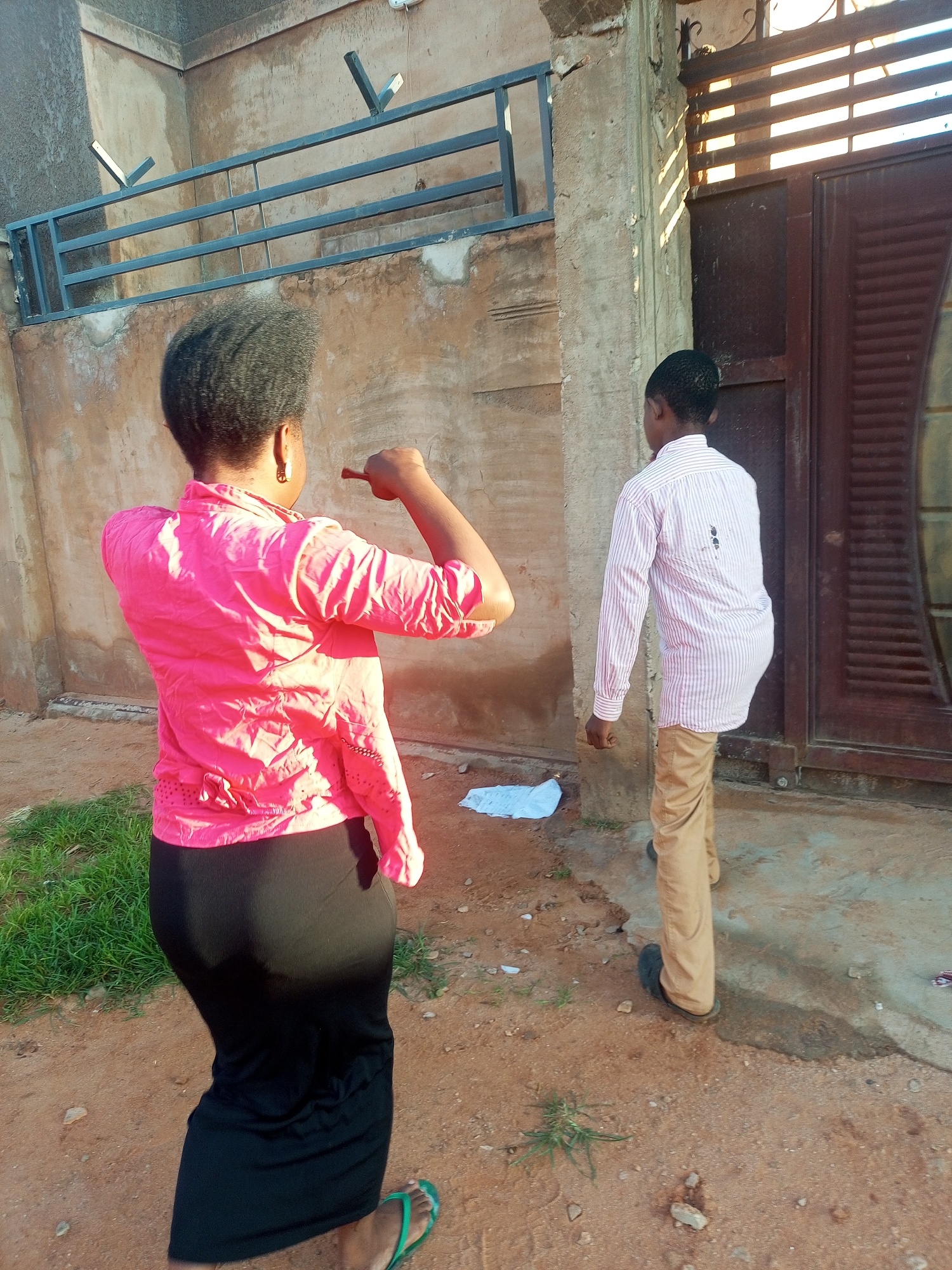 The image depicts two individuals in what appears to be an outdoor urban environment in Tempure, Nigeria. A woman, viewed from behind and to the side, is wearing a vibrant pink collared shirt and a long black skirt. Her hair is styled in a short, natural afro, and she is wearing gold hoop earrings. She is raising her right arm and appears to be pointing or gesturing with her index finger, which is extended. In front of her and to her right, a young male is also viewed from behind. He is wearing a light-colored, pinstriped long-sleeved shirt and tan trousers. His stance suggests he is walking or standing near a large, brown, slatted metal gate. The ground is a mixture of dirt and concrete, with some sparse green vegetation growing in patches. A wall with a metal railing is visible in the background, and the overall impression is of a utilitarian, possibly residential, area in Nigeria.
