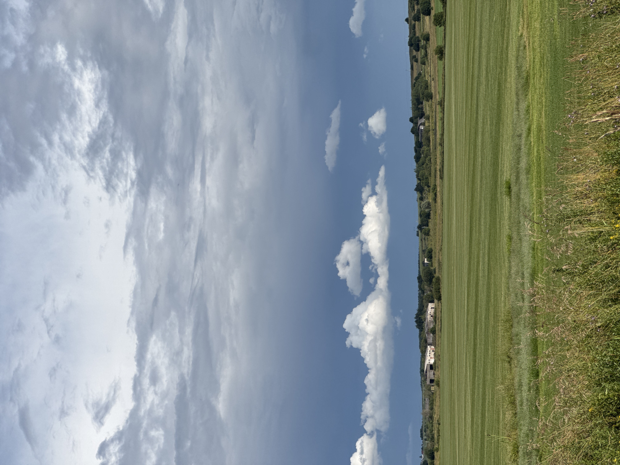 This image depicts a rural landscape in Kempelevo, Russia, under a partly cloudy sky. The scene is dominated by a vast, meticulously mown green field that stretches horizontally across the frame, marked by the characteristic stripes of a lawnmower. To the left of the field, a line of trees and some buildings are visible, suggesting a small settlement or farmstead nestled amidst the countryside. The sky is a mix of blue and gray, with streaks of white clouds. The overall impression is one of a peaceful, agricultural setting on what appears to be a clear day. No people or animals are visible in the frame. The image seems to have been taken during daylight hours, possibly late morning or early afternoon.