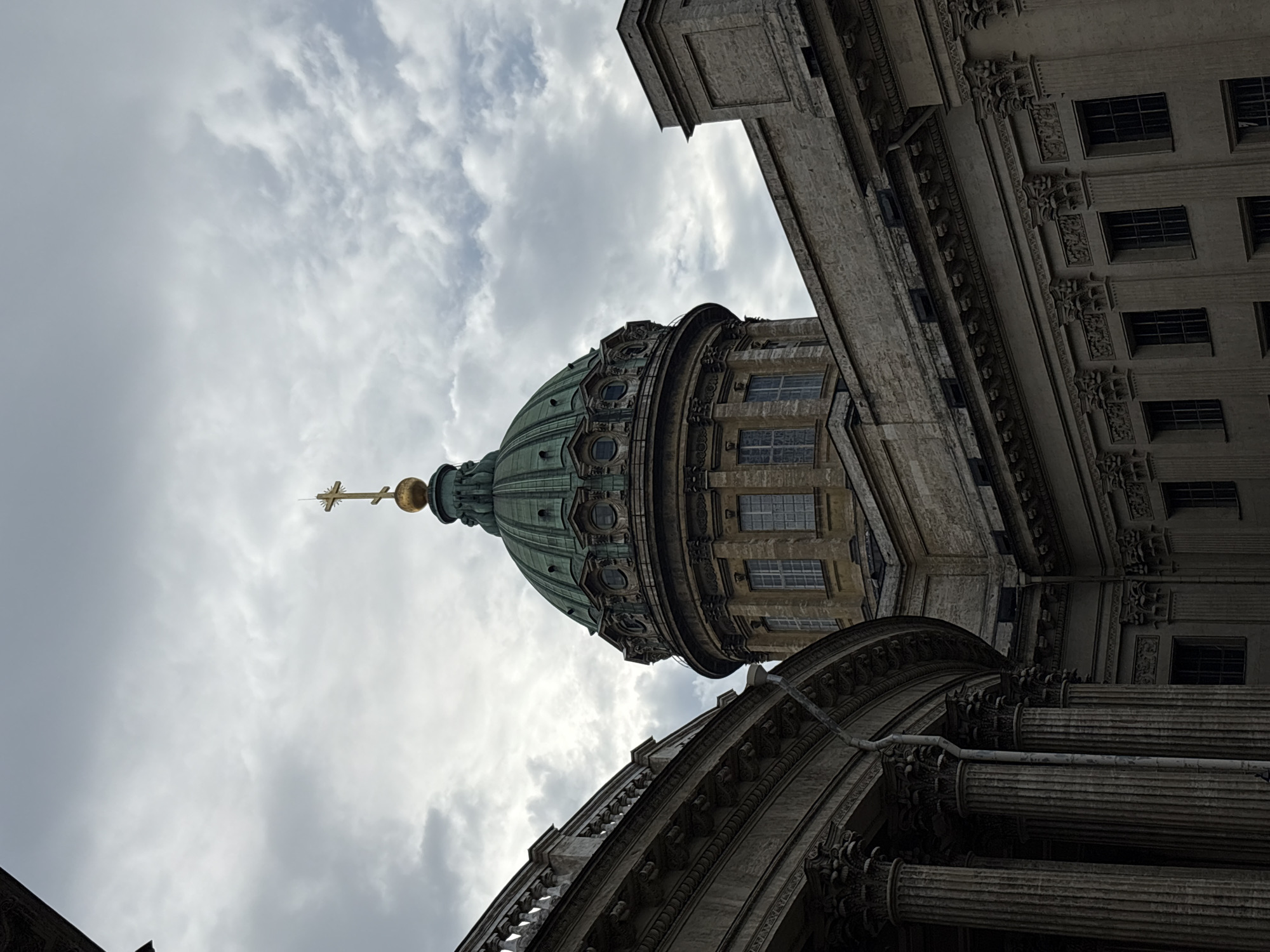 The image shows a view of Kazan Cathedral in Saint Petersburg, Russia, from a low angle, looking upwards. The cathedral's prominent green dome, topped with a golden cross, dominates the upper central portion of the frame. The dome is surrounded by smaller domes and decorative elements. The architecture of the cathedral is classical, with visible columns, windows, and intricate detailing on the façade. To the left of the dome, a curved colonnade with ornate capitals is partially visible, forming a grand entrance or wing of the building. The sky above is overcast with dramatic clouds, casting a diffused light on the scene. The overall mood is one of grandeur and solemnity.