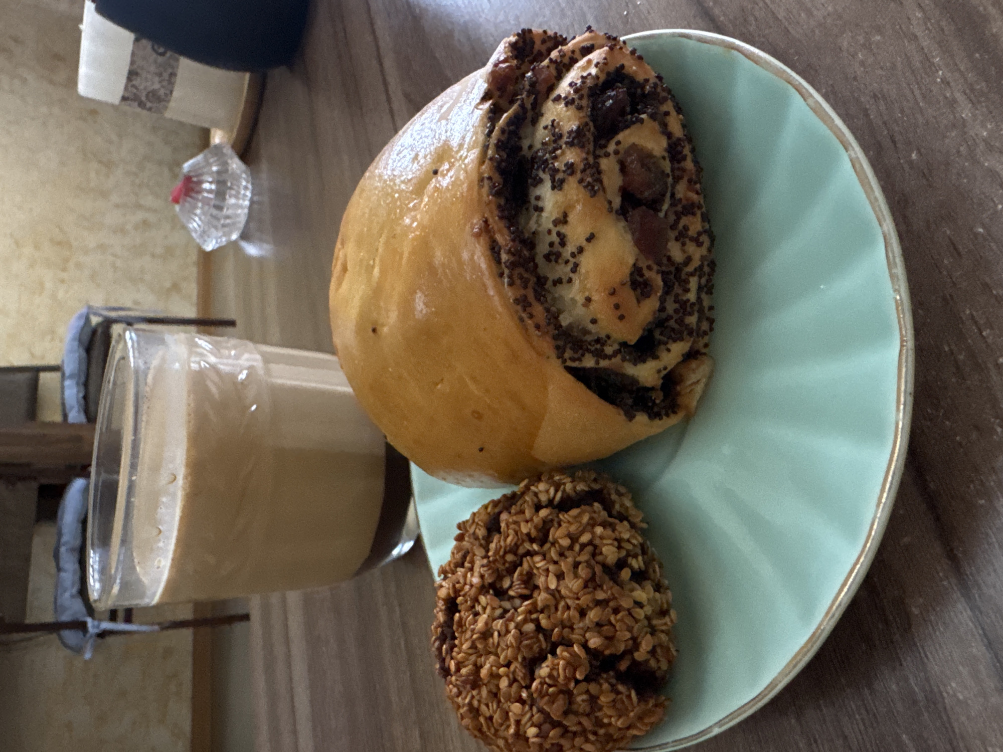 On a wooden surface is a light blue plate with a gold trim, upon which sits a pastry roll filled with poppy seeds and raisins, and a cookie covered in sesame seeds. Next to the plate is a clear glass filled with a light brown liquid, likely a beverage. The scene is indoors, the specific location and city unknown. No people are visible.