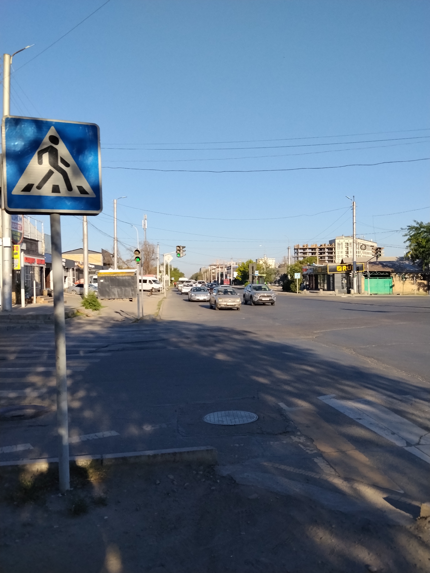 The image shows a street scene in Bishkek City, Kyrgyzstan. In the foreground, a pedestrian crossing sign is visible on the left side of the image. The sign is blue and white, with a silhouette of a person walking across a crosswalk. A traffic light is visible in the middle ground, showing a green light. Several cars are driving on the road. The background shows buildings and trees. The sky is clear and blue. The overall scene depicts a typical urban environment on a sunny day.