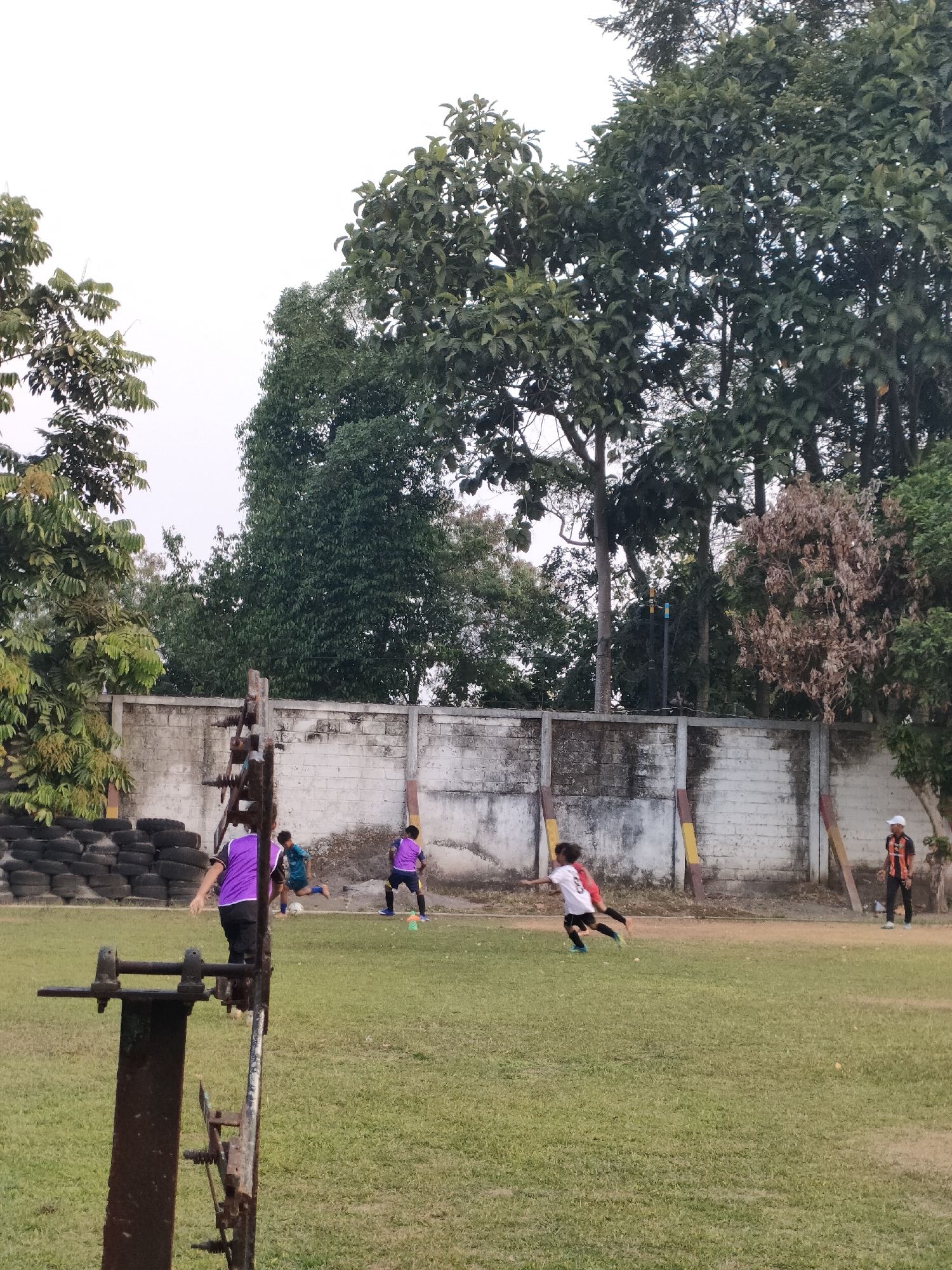 This image captures an outdoor scene at a grassy field, likely a sports ground, with a white brick wall in the background. The wall is adorned with colorful vertical posts, red and yellow, at intervals. To the left of the wall, a stack of used tires is visible. A group of individuals, appearing to be children, are engaged in what looks like a soccer game. One child in a purple jersey is actively participating, while another in a white jersey with black shorts is running. Further to the right, another child in a purple jersey is standing, and a person in an orange and black shirt with a white cap is observing. The environment is characterized by lush green grass and mature trees in the background, suggesting a location within a park or open space in the city of Banguntapan, Indonesia. The overall atmosphere is one of casual sporting activity.