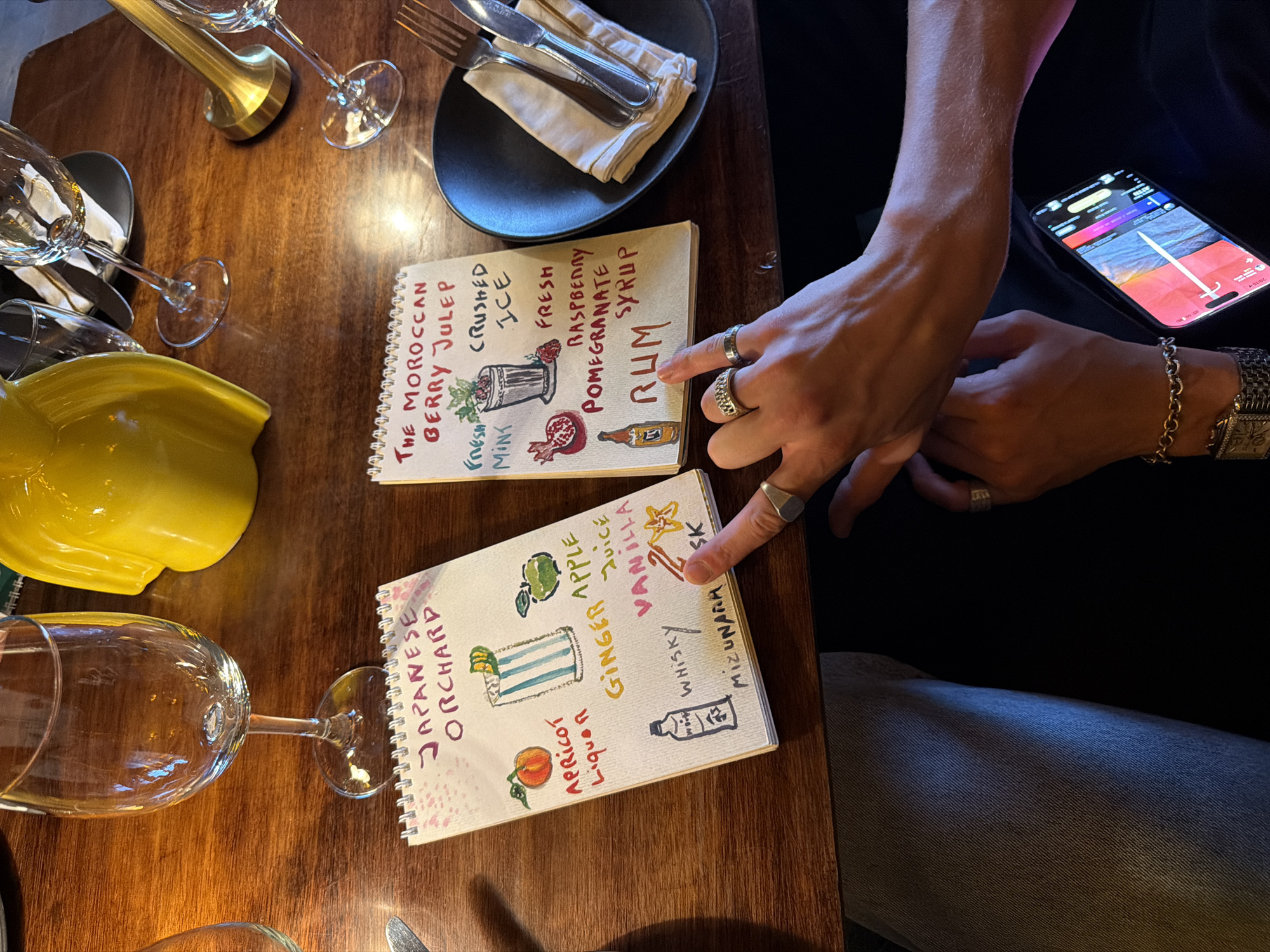The image depicts a close-up view of a wooden table in Marrakesh, Morocco, where two handwritten cocktail menus are laid out. The table is set with a wine glass, a yellow pitcher, and silverware on a black plate.
The left menu, titled "JAPANESE ORCHARD," is illustrated with a drawing of an apricot, a cocktail glass, and an apple. The ingredients listed include "APRICOT LIQUOR," "GINGER APPLE JUICE," "VANILLA," "WHISKY," and "MIZUNARA."
The right menu, titled "THE MOROCCAN BERRY JULEP," features drawings of mint, crushed ice, a pomegranate, a raspberry, and a bottle of rum. The ingredients listed are "FRESH MINT," "CRUSHED ICE," "FRESH RASPBERRY POMEGRANATE SYRUP," and "RUM."
Two individuals' hands are visible in the foreground. One hand, adorned with rings, is pointing to the "RUM" on the right menu. The other hand, with a bracelet and watch, is resting near a smartphone displaying a colorful interface. The overall scene suggests a relaxed setting, possibly a restaurant or bar, where patrons are deciding on drinks. The presence of a yellow pitcher might indicate it's being used for serving drinks or is a decorative item. The background shows more tableware and a glimpse of the ambient environment.