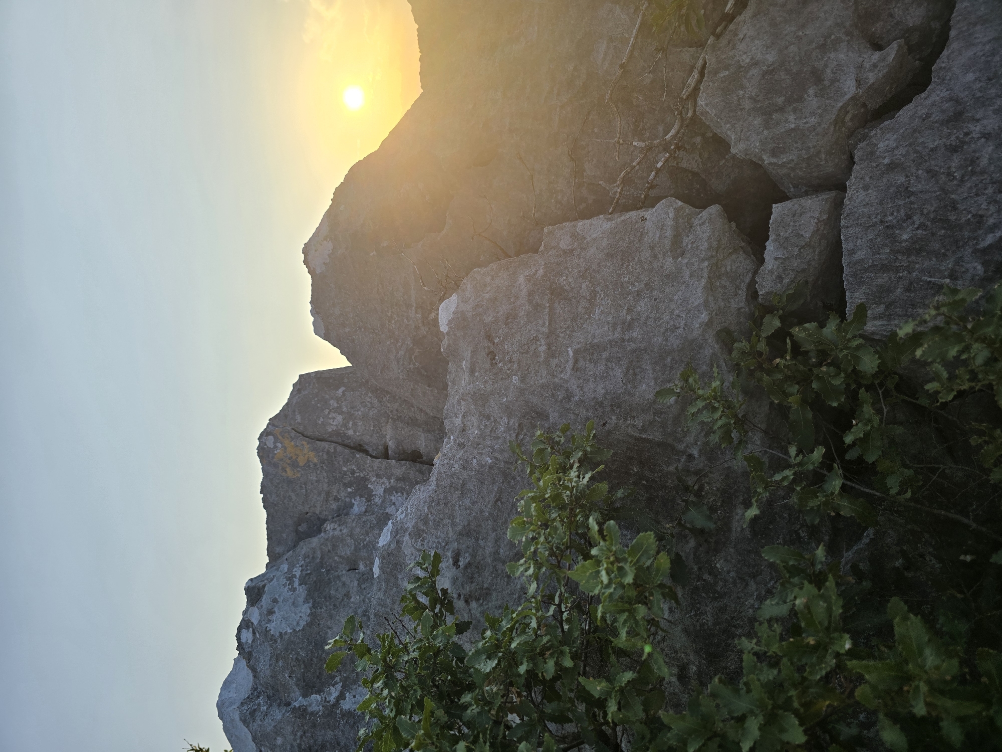 The image depicts a natural landscape featuring large, gray rock formations illuminated by the setting sun. The sun, a bright orb, is positioned in the upper right quadrant of the image, casting a warm glow and creating lens flare. Green foliage, likely from a bush or small tree, is visible in the lower left and along the bottom edge of the frame, contrasting with the stony textures of the rocks. The sky above is a soft gradient of pale blue and yellow, indicating the time of day is twilight. The rocks are rough and irregular, with some appearing weathered and possibly marked with yellow paint, suggesting a trail or directional indicator. The overall scene is one of a rugged, outdoor environment, possibly a hiking trail or a mountainous area, with a serene sunset as the backdrop. The location is specified as Podgorica, Montenegro.