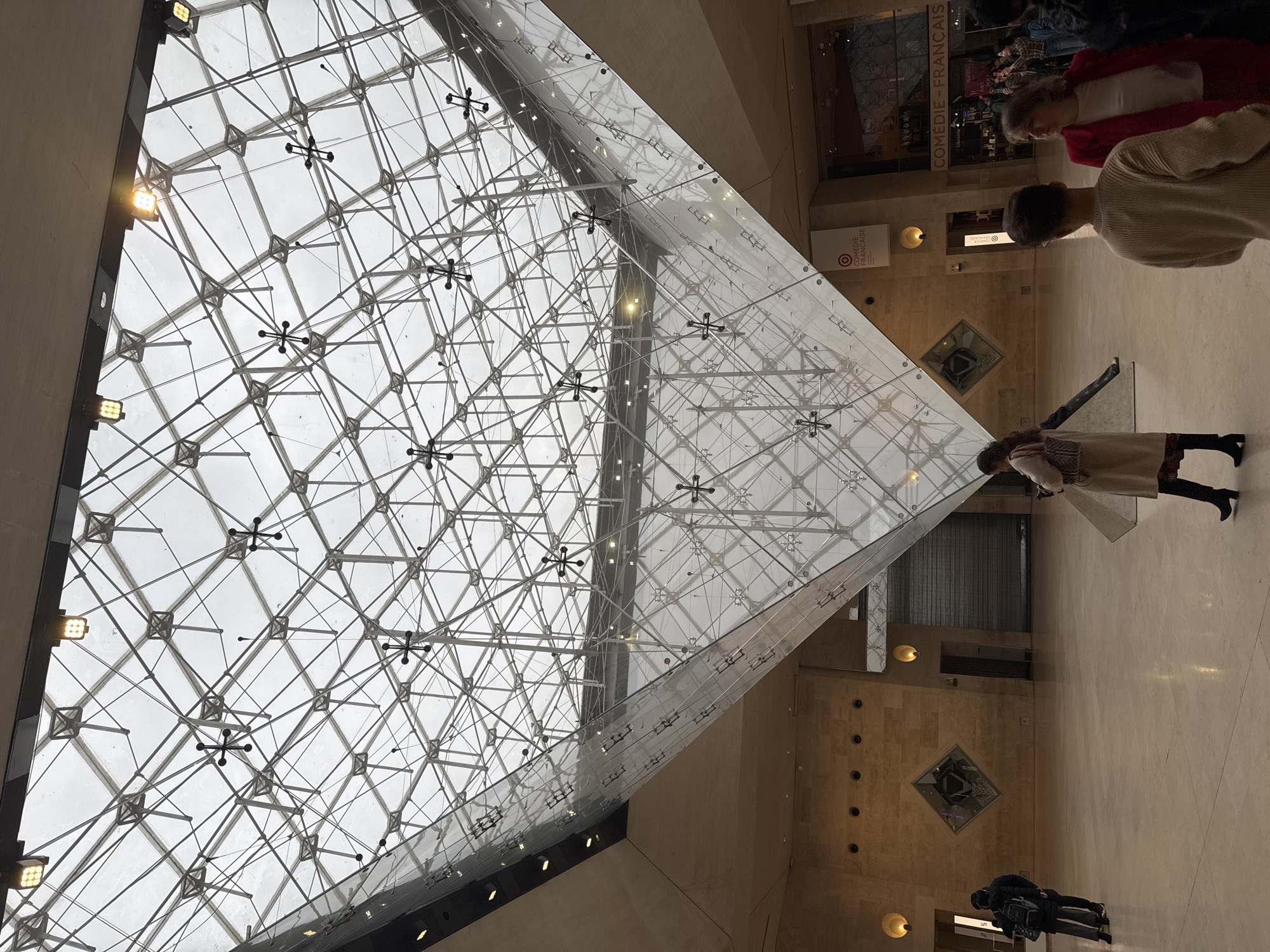 The image shows the interior of the Louvre Museum in Paris, France, focusing on the glass pyramid structure. The pyramid is composed of a geometric framework of metal beams and glass panels, ascending towards a cloudy sky visible through the glass. Several people are within the frame: two standing near the base of the pyramid, another pair near an entrance, and two additional people in the distance. The museum's interior features light-colored marble or stone flooring and walls, and includes architectural details such as recessed lighting and diamond-shaped insets in the wall. Signage for "COMÉDIE - FRANÇAISE" is also visible.
