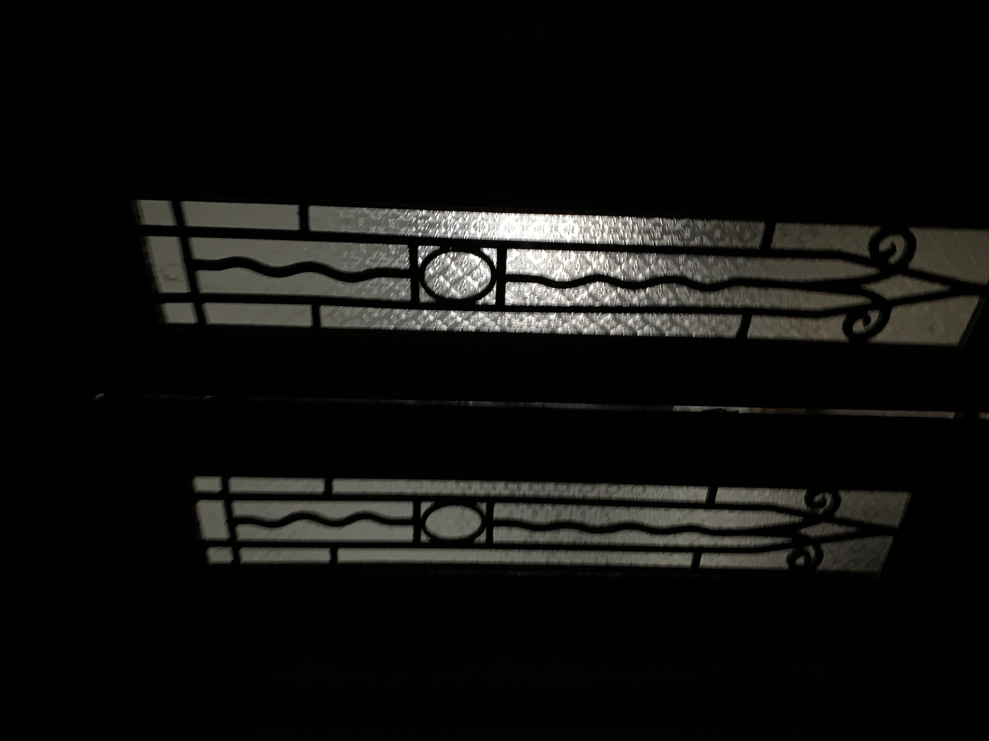 The image shows two horizontal decorative glass panels set within a dark framework. The panels feature a repeating pattern of straight lines forming rectangular segments, a central wavy line, a circle intersecting the wavy line, and decorative swirls at each end. The light source appears to be behind the panels, illuminating them against the dark surroundings. It's impossible to determine the location in Tel-Aviv, Israel, or to provide any information about people, subjects, actions, interactions, or other environmental details beyond what is visible in the image.