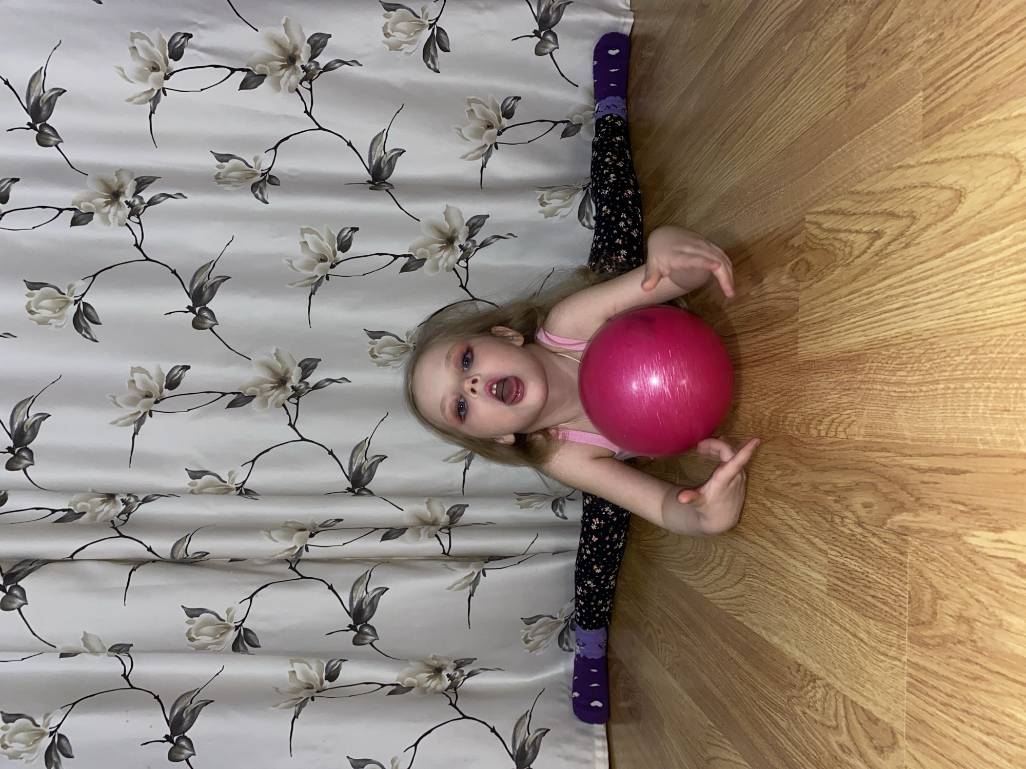 A young girl, approximately 4-5 years old with blonde hair and blue eyes, is lying in a split on the floor between a patterned curtain and a light wood-colored laminate floor, holding a red ball. She wears a pink tank top and dark leggings with a small floral pattern, ending in purple socks with a small design. Her mouth is open, and she appears to be vocalizing. The curtain has a floral pattern consisting of large, pale blossoms and dark grey stems. The image appears to have been taken indoors. There is no indication of this image being taken in Votkinsk, Russia.