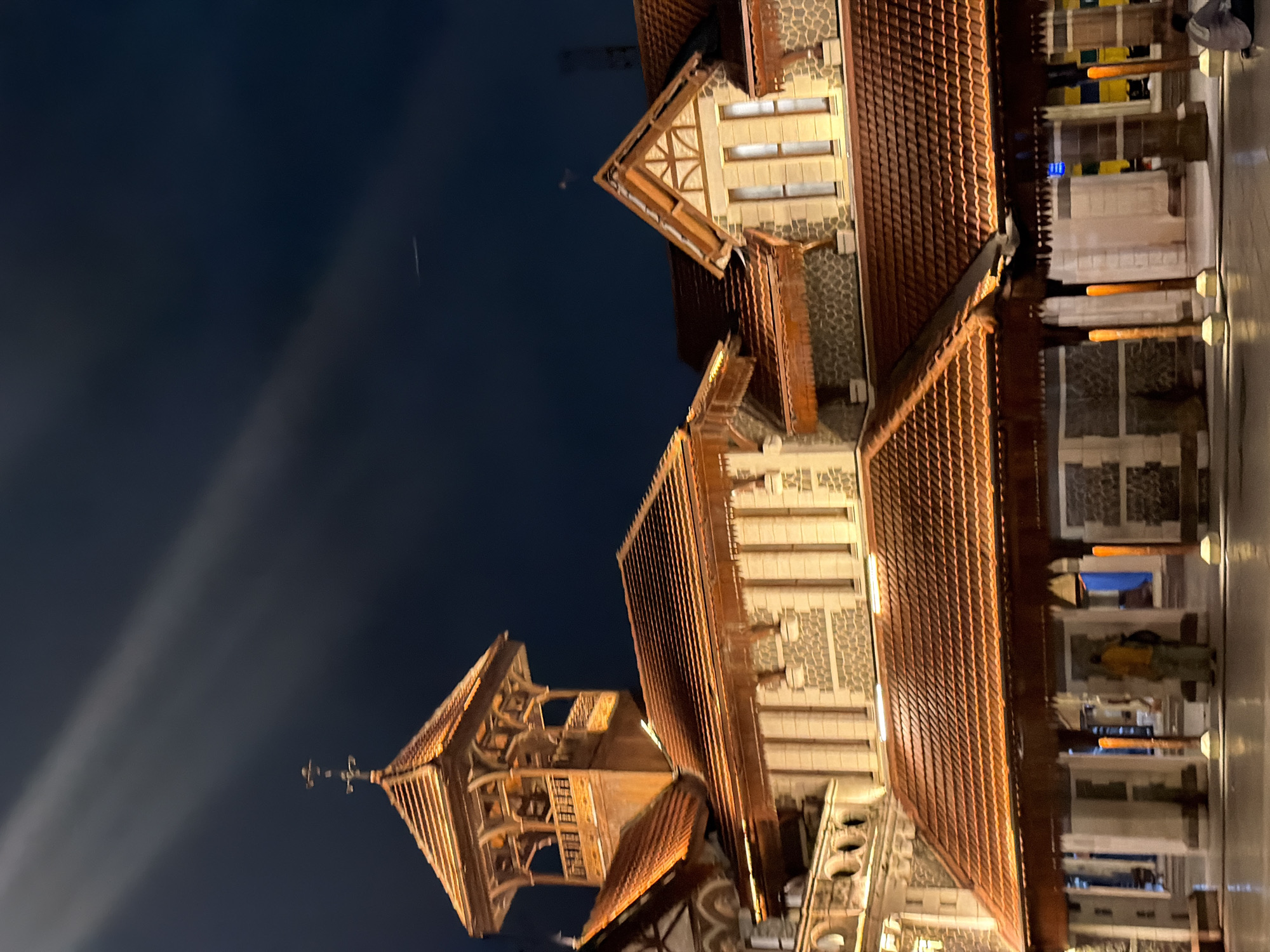 This is a nighttime photograph of the Taj Mahal Palace Hotel in Mumbai, India. The iconic building's distinctive red-tiled roofs and stone facade are illuminated by warm artificial lighting. A prominent tower with intricate wooden latticework stands tall against the dark night sky. Rays of light streak across the sky, possibly from spotlights or reflections. In the foreground, a person in a yellow shirt and khaki pants is partially visible standing under a covered walkway. Another person is seated on the ground to the right. The ground appears wet, suggesting recent rain or a reflective surface. The overall impression is one of grandeur and architectural splendor captured at night.
