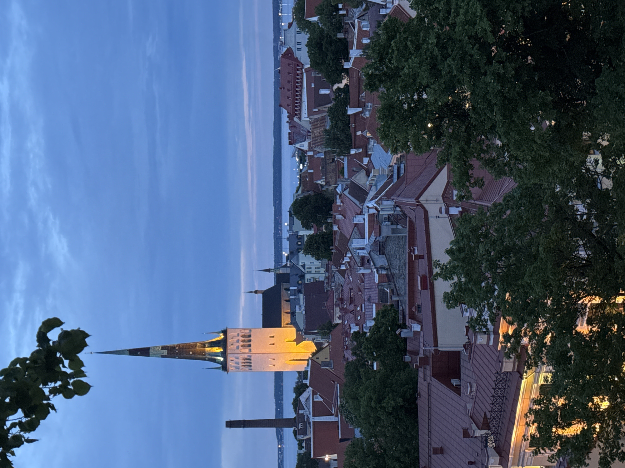 The image captures an aerial view of Tallinn, Estonia, at dusk. The iconic St. Olaf's Church, with its tall, illuminated spire, dominates the skyline. The church's white facade is bathed in warm yellow light, highlighting its architectural details. The spire is a striking element, tapering to a point and adorned with decorative bands of darker color.
Below and around the church, a dense collection of buildings with red tiled roofs forms a picturesque urban landscape. The rooftops vary in shape and size, indicating a mix of architectural styles and ages. Numerous chimneys punctuate the roofs. Green trees, with lush foliage, are interspersed throughout the cityscape, adding a natural element to the scene.
In the background, a body of water, likely the Baltic Sea, stretches towards the horizon. A hint of a distant shoreline or islands can be seen across the water, and a few lights twinkle on the far side. The sky is a deep blue, transitioning from a lighter hue near the horizon, with scattered clouds contributing to the twilight atmosphere. To the left of the church, a tall, dark brick chimney stands as a stark industrial element against the softer architectural and natural features. The overall scene evokes a sense of historic charm and tranquility as day transitions into night.