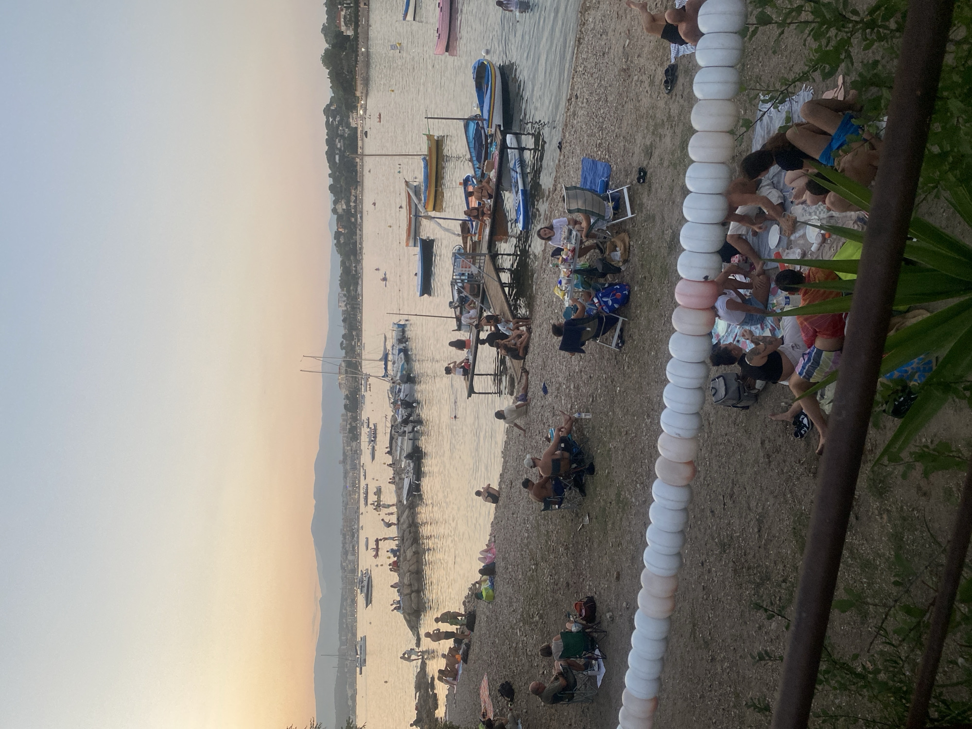 The image depicts a lively scene at a beach in Antibes, France, during what appears to be sunset. The foreground is occupied by a gravelly beach with several groups of people engaged in various activities. One group is having a picnic on blankets, with food and drinks spread out. Another group is seated in portable chairs, observing the surroundings. Further down the beach, individuals are relaxing on towels and sitting on the pebbles.
In the mid-ground, a wooden jetty extends into the calm, sparkling water, with several small boats and a paddleboard moored nearby. People are seen sitting on the jetty, some interacting with those in the water. The water is populated with more boats of various sizes, including sailboats and motorboats, anchored or moving slowly. Some individuals are swimming or wading in the water.
The background reveals the coastline of Antibes, with a dense collection of buildings and lush green hills rising behind them. The sky is a soft gradient of orange, yellow, and blue, indicative of the setting sun, casting a warm glow over the entire scene. The overall atmosphere is one of leisure and enjoyment, with people taking advantage of the beautiful coastal setting.