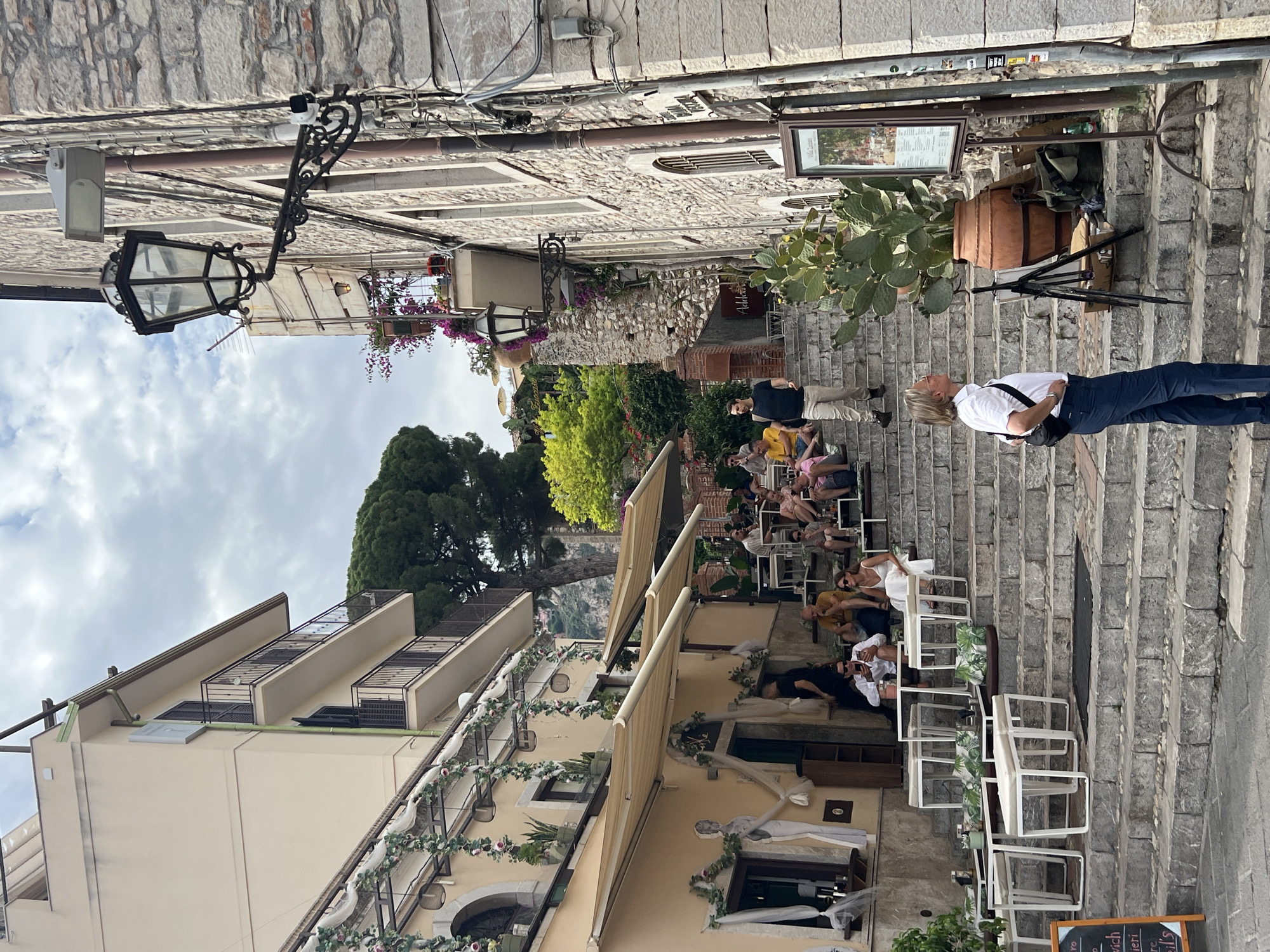 This image depicts a steep, stone staircase lined with tables and chairs for outdoor dining, characteristic of the city of Taormina, Italy, known for its tiered architecture and vibrant street life. On the left, a building with cream-colored stucco walls features awnings extended over tables and chairs set up on a lower level, decorated with flowing white fabric and floral garlands. Several people are seated at these tables, appearing to be dining or relaxing.
The staircase itself is a central feature, ascending upwards and populated with more tables and chairs. People are seated on various steps, some engaged in conversation or looking at their phones. A man in a white shirt and dark pants stands at the bottom of the staircase, facing up and to the right, holding a camera bag slung across his body. To his right, a large terra cotta pot holds a prickly pear cactus, and behind it, a tripod with art supplies suggests an artist might be working in the vicinity. A menu board stands near the cactus.
Further up the stairs, more individuals are seated, and a man in a black t-shirt and light-colored pants walks down. The architecture on the right side of the image is made of rough-hewn stone, with windows and balconies. Ornate streetlights are mounted on the building. In the background, lush green trees and vegetation add to the scenic setting, hinting at the hilly terrain of Taormina. The sky is overcast, suggesting a possibly temperate day. The overall impression is one of a bustling, picturesque European street scene with a focus on outdoor leisure and dining.
