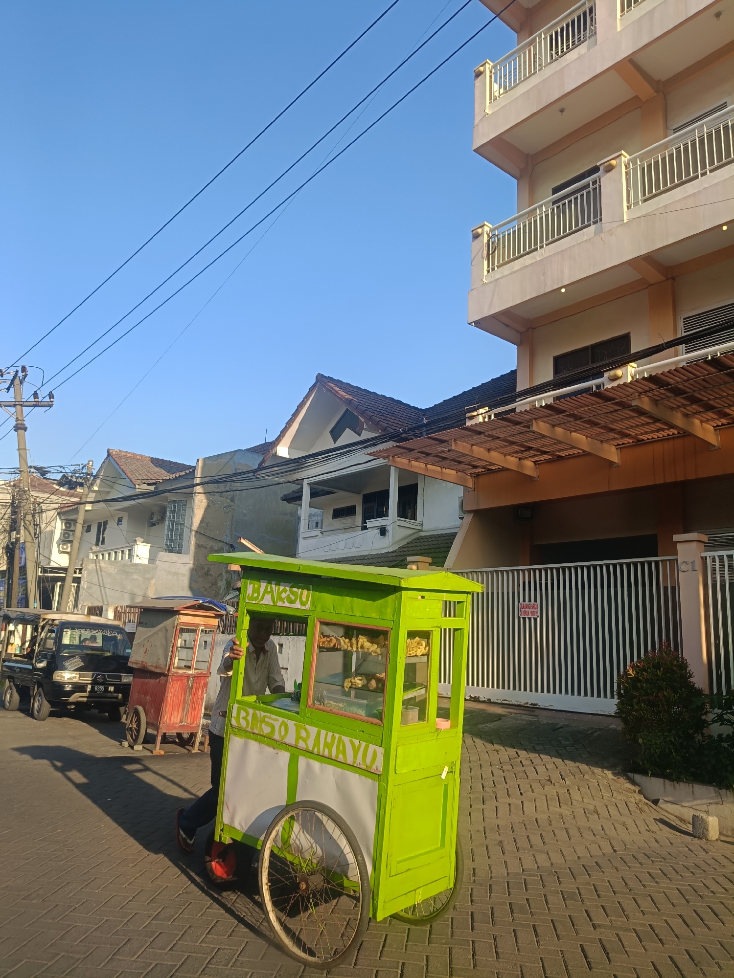 In Surabaya, Indonesia, a street scene unfolds under a clear blue sky. A bright green food cart, adorned with "BAKSO RAHAYU" text, is positioned on a paved walkway. A person, wearing a light-colored collared shirt and dark pants, is standing beside the cart. Behind the cart, a red and white food cart is visible, followed by a black truck. In the background, several buildings are present, including a modern multi-story building with balconies and other residential structures. Electrical wires crisscross the sky, and the sun casts long shadows on the ground, indicating it is likely late afternoon.