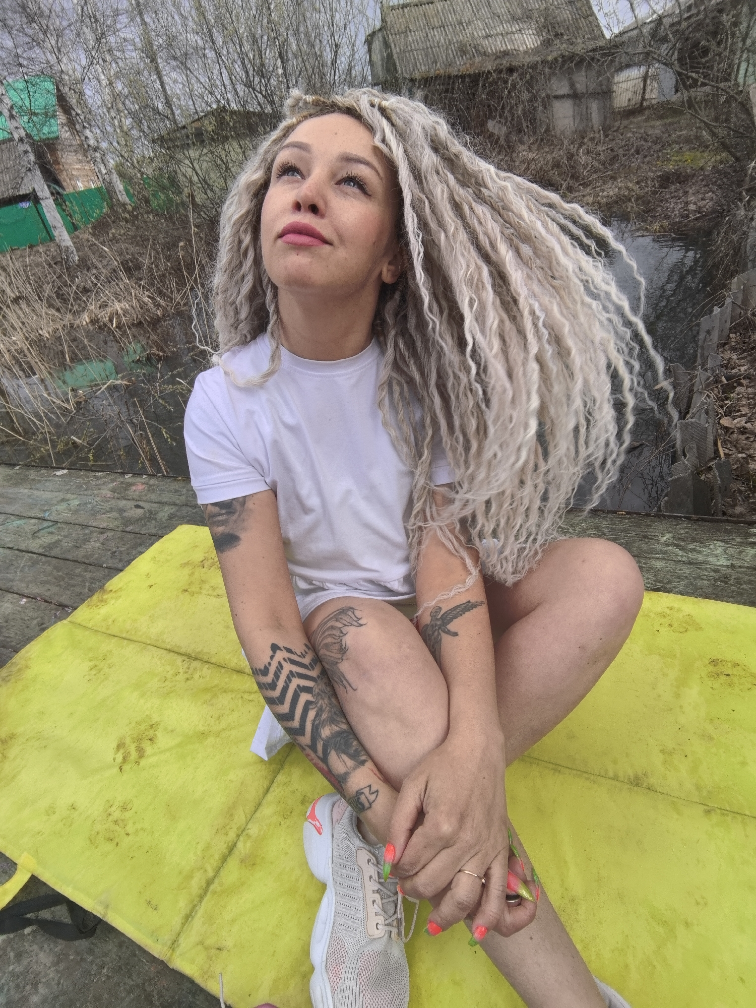 A young woman with long, curly, light blonde dreadlocks sits cross-legged on a bright yellow mat. She is wearing a simple white t-shirt, light-colored sneakers with pink accents, and has several tattoos on her arms and legs, including geometric patterns and an avian design. Her fingernails are painted in a striking neon orange and green. She looks upwards with a thoughtful or contemplative expression, her lips painted a soft pink.
The setting appears to be an outdoor, somewhat rustic or rural location in Noviy, Russia, during daytime under an overcast sky. The yellow mat rests on a weathered wooden surface, possibly a pier or deck. In the background, there is a small body of dark water or a murky stream, surrounded by dry reeds, bare trees, and withered vegetation. Further back, old, dilapidated wooden structures with corrugated roofs are visible, along with hints of green-painted walls on a smaller building or shed. The overall scene suggests a quiet, possibly neglected area.