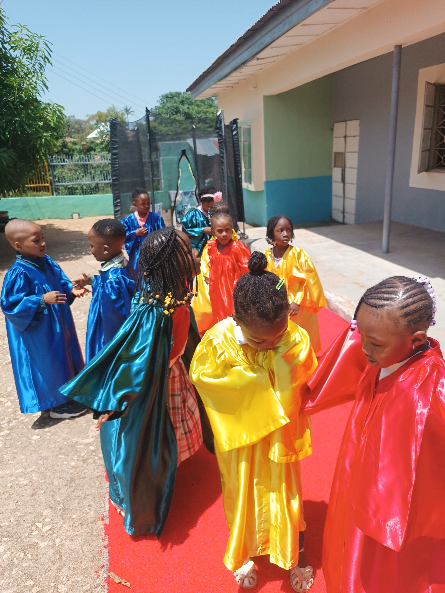 In Jos, Nigeria, a group of children, dressed in vibrant robes of blue, green, yellow, and red, are gathered outdoors. The children appear to be participating in some form of performance or ceremony, perhaps a school event, as they are arranged on a red carpet. Some are looking at each other, possibly interacting or waiting for their turn, while others gaze forward. The setting suggests a school or community center, with a building featuring a light-colored exterior, windows, and a corrugated roof visible in the background. A trampoline is also present, adding to the playground-like atmosphere. The bright sunlight indicates it is daytime, and the clear sky suggests fair weather. The children's attire and the celebratory setup imply a joyful and organized occasion.