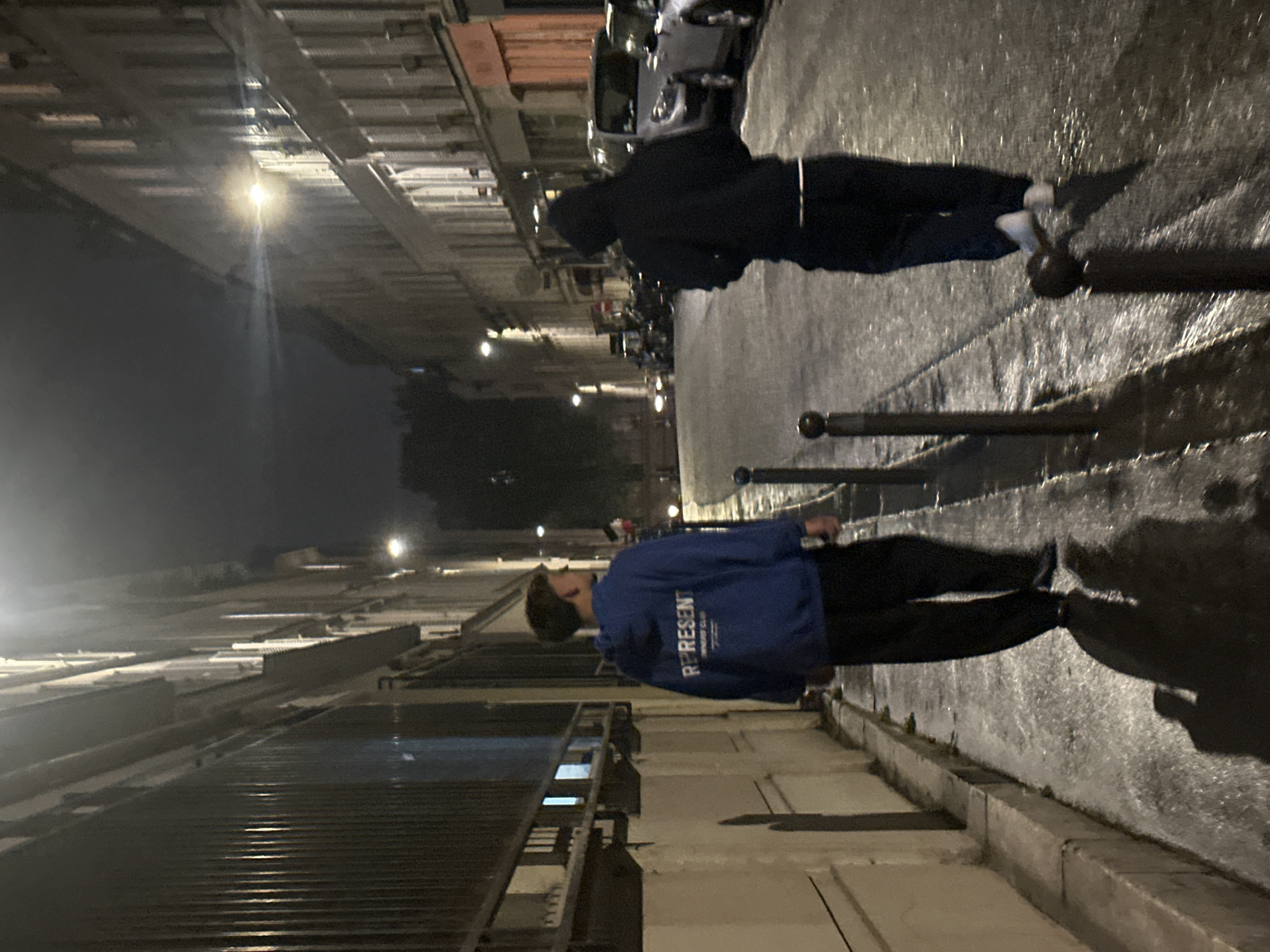 Two individuals walk on a wet Parisian street at night. One wears a blue "Represent" jacket, and the other wears a dark hooded sweatshirt. Streetlights illuminate the scene, reflecting off the wet pavement. Buildings line the street, with some covered by metal shutters or scaffolding. Cars are parked along the curb. Bollards are visible along the sidewalk.