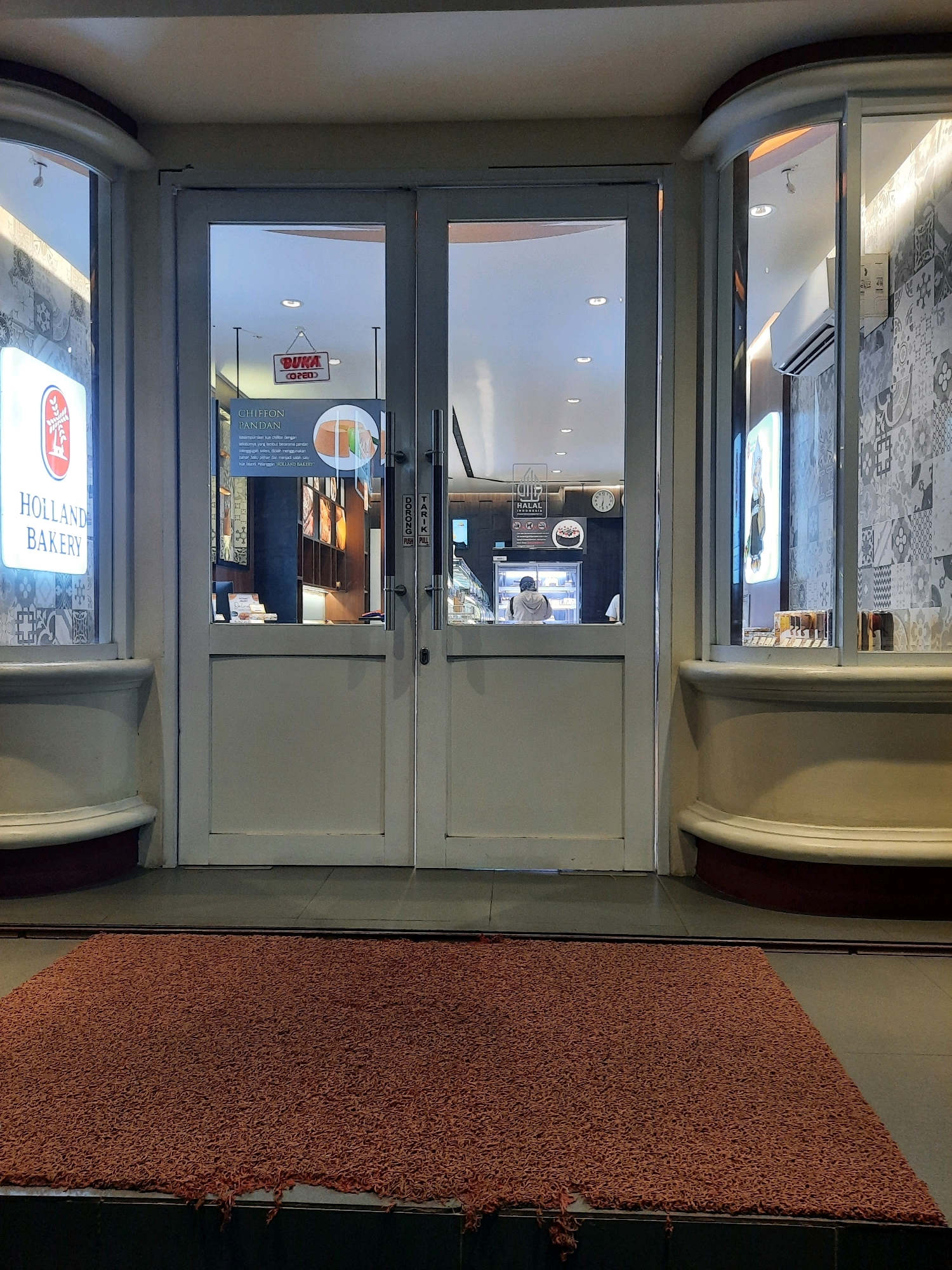 The image shows the entrance to a Holland Bakery in East Jakarta, Indonesia. The facade features double glass doors with white frames. A red doormat is placed on the floor in front of the entrance. To the left of the doors, a lit sign with "HOLLAND BAKERY" is visible. Through the glass doors, the interior of the bakery is dimly lit, showing display cases and shelves. A "BUKA OPEN" sign is hanging on the inside of the left door. On the right door, a "TARIK PULL" sign is visible. The architecture includes curved decorative elements on the exterior walls.