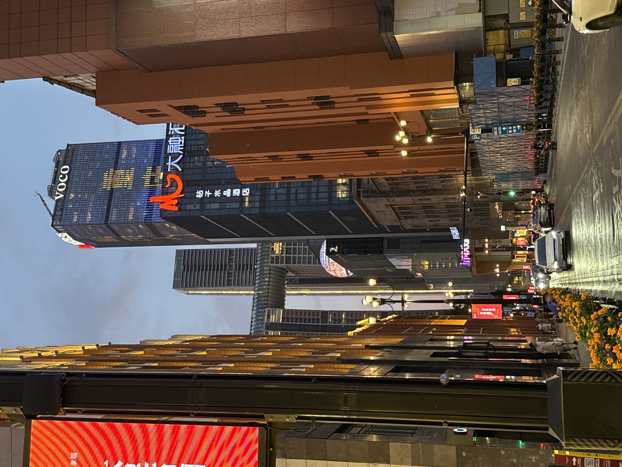 This is a vertical, eye-level, street-level shot of a bustling urban scene at dusk in Yuzhong District, China. Tall, modern buildings with illuminated signage dominate the skyline. The building on the left features a prominent "VOCO" logo in blue lights and Chinese characters that appear to say "大慈寺" (Dàcí Sì, Great Mercy Temple) in red and white. Another building displays "HOTEL INDIGO" in white text. In the foreground, a bright red LED screen with a striped pattern is visible.
The street is wet, suggesting recent rain, and reflects the glowing lights of the city. Cars are visible on the road, indicating moderate traffic. Several street lamps with multiple bulbs illuminate the sidewalk and the buildings. Orange flowers in planters line the street, adding a touch of color to the urban landscape.
A few people are seen walking along the sidewalk, some appearing to be dressed in light-colored clothing, suggesting they are pedestrians. The lighting indicates it is either dusk or early evening, with artificial lights taking over from the fading daylight. The overall atmosphere is that of a lively commercial district at the end of the day. Construction scaffolding is visible on one of the buildings, suggesting ongoing development or renovation. Signs with blue arrows and Chinese characters are also present, providing directional information.