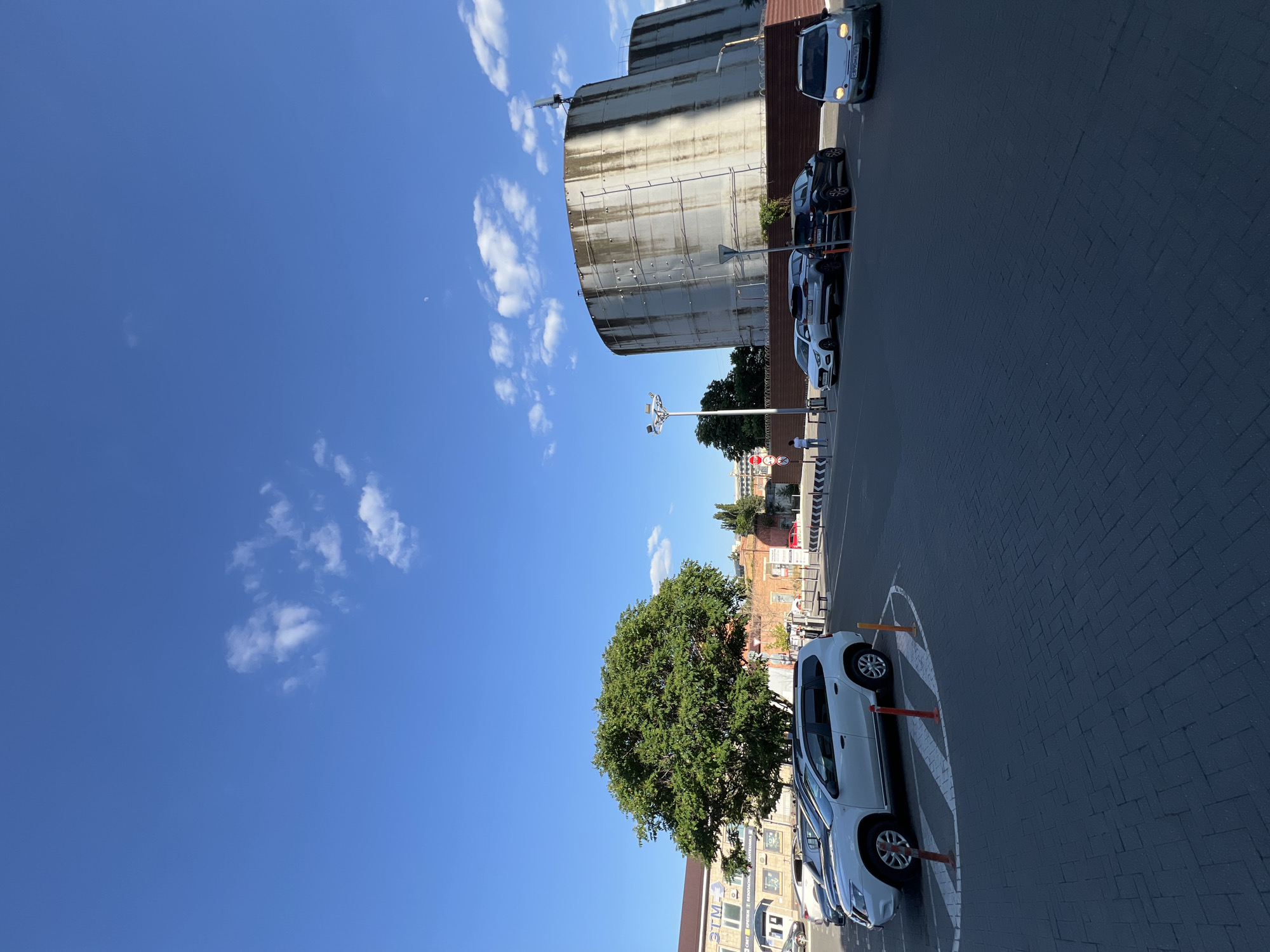 This media file depicts an outdoor scene under a clear blue sky with scattered white clouds, suggesting a daytime setting. The foreground is dominated by a paved parking area, laid with dark grey bricks in a herringbone pattern. Several cars are parked in a line on the left side of the frame. A white SUV, facing away from the camera, is the most prominent vehicle, positioned within a marked parking bay with orange bollards. To its left, a row of other cars, including darker sedans and a white hatchback, are visible.
In the mid-ground, a large, mature green tree with a dense canopy stands out. Behind the tree, a multi-story building with a light-colored facade and numerous windows is partially visible, along with some red-brick structures. Further in the background, more buildings and some infrastructure can be seen.
To the left of the cars and tree, a massive, cylindrical concrete structure with a weathered, stained surface occupies a significant portion of the frame. The structure appears industrial or utilitarian. A metal antenna or tower is attached to its top.
The overall scene suggests a parking lot or an area adjacent to industrial or commercial buildings in Krasnodar, Russia. The weather appears to be pleasant and sunny. There is no discernible activity or event taking place, with the image capturing a static moment. Some faint text is visible on the license plates of the cars, but it is not clear enough to be legible. A street sign with multiple circular signals and a directional arrow is also present near the tree.