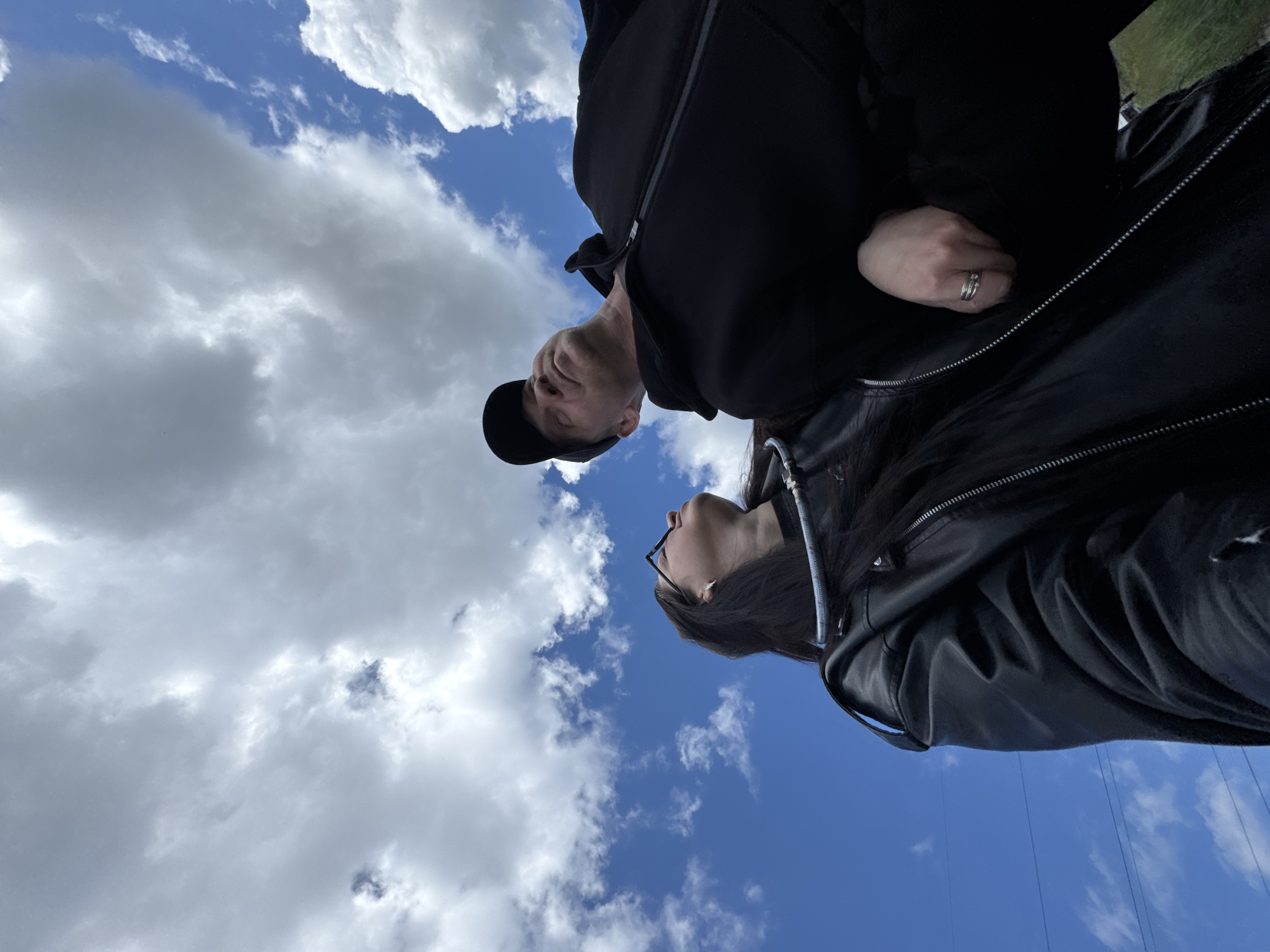 The image presents a low-angle view of two individuals, a man and a woman, looking upwards towards a bright blue sky partially covered by white and gray clouds. The perspective is from directly below the subjects, who are positioned close to each other.
The man, on the left, is seen in profile, wearing a dark baseball cap and a dark jacket or hoodie. He has fair skin and visible stubble or a short beard, with his gaze directed intently upwards. To his right, the woman, also in profile, has long dark hair and wears glasses. She is dressed in a dark leather jacket, and her right hand, adorned with several rings on her ring finger, is visible against her clothing.
The dominant feature of the scene is the expansive sky, characterized by patches of clear blue interspersed with large, fluffy cumulus clouds. The weather appears fair and bright, indicating it is daytime. No specific location cues are discernable beyond the sky, though faint lines, possibly wires or ropes, are visible against the blue sky in the bottom right corner, suggesting an open outdoor environment, perhaps near some infrastructure. Both individuals appear calm and focused, with their expressions conveying contemplation or observation of something above them. No text is visible in the image.