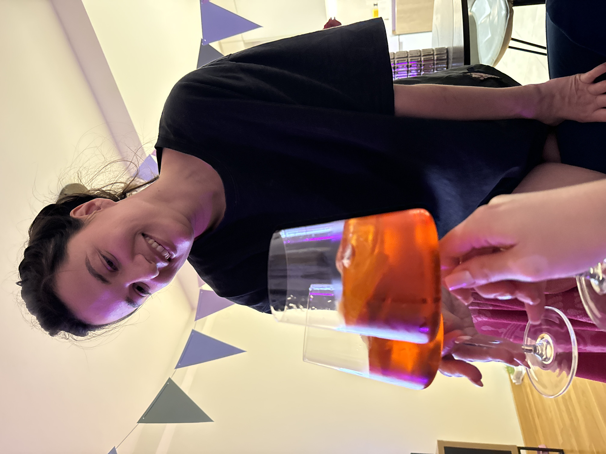 A woman with dark hair pulled back, wearing a black garment, smiles while holding a glass filled with an orange liquid. Another person's hand is also holding a glass, presumably filled with the same drink. The background includes a wall decorated with triangular pennants. The location is indoors. The city of Belgrade, Serbia is not discernible from this image.