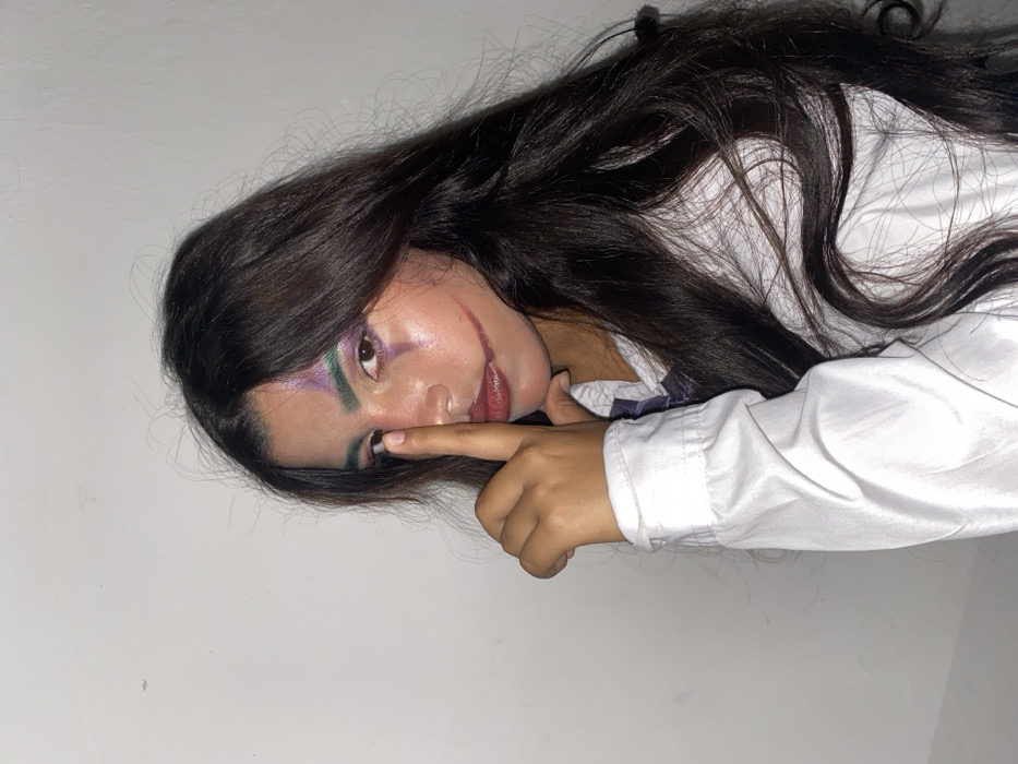 The image, oriented on its side, features a person with long, dark hair, wearing elaborate makeup and a white collared shirt. The person is positioned against a plain, light-colored wall or surface, possibly lying down or leaning back, and looking directly towards the viewer.
The central focus is the person's face, adorned with striking, colorful makeup. Vibrant purple eyeshadow is blended with green on the eyelids and extends upwards, creating a dramatic effect, particularly around the left eye (from the viewer's perspective). A distinct dark green line or streak is painted on or just above the left eyebrow. The lips are colored with a reddish-pink lipstick or tint. A prominent detail is the red or dark red lines drawn from the corners of the mouth, extending upwards and outwards onto the cheeks, resembling a "Joker smile" or a "Glasgow smile." The skin appears smooth with some highlights, suggesting a glow or intentional highlighting makeup.
The person's right index finger is gently touching the area just below their left eye, near the cheekbone, a pose that draws attention to the eye makeup and the overall facial artistry. Their long, dark brown or black hair is wavy and flows outward, spreading across the light background behind their head and shoulder.
They are wearing a white button-up shirt with a collar. A hint of a darker garment or accessory, possibly a tie or ribbon, is visible near the neck and collar area of the shirt. The lighting appears soft and even, highlighting the features without harsh shadows, suggesting an indoor setting. No text is visible in the image. The overall impression is that of a creative or costume-themed portrait.