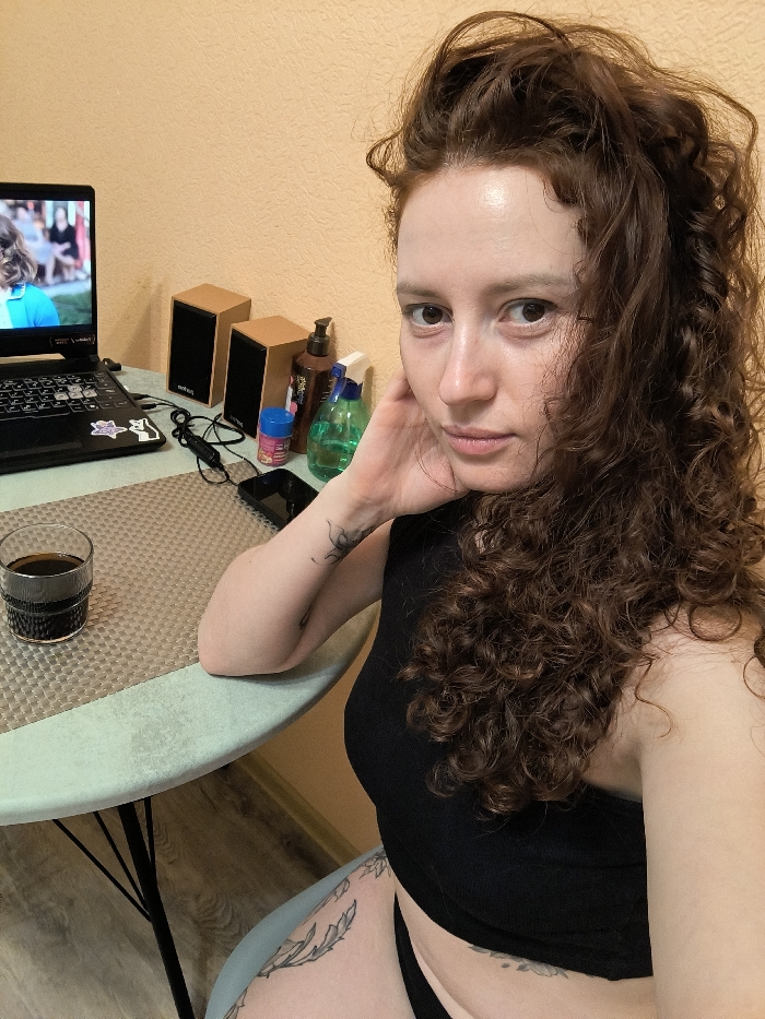 A female individual with long, curly dark reddish-brown hair is depicted seated at a light mint green round table in an indoor setting in Saint Petersburg, Russia. She wears a black sleeveless crop top and black underwear, revealing tattoos on her left forearm and left upper thigh. Her right hand rests near her cheek, while her left forearm is positioned on the table. She gazes directly into the camera. On the table to her left, a clear glass filled with a dark liquid sits on a grey-patterned placemat. A black open laptop, displaying a video of a person, is positioned next to the glass. Behind the laptop are two small, light wood-grain speakers. Further to the right on the table are a black cable with an in-line control, a small black rectangular device, a brown pump bottle, a blue container, and a green transparent spray bottle. The background consists of a plain light peach-colored wall and a wooden floor.