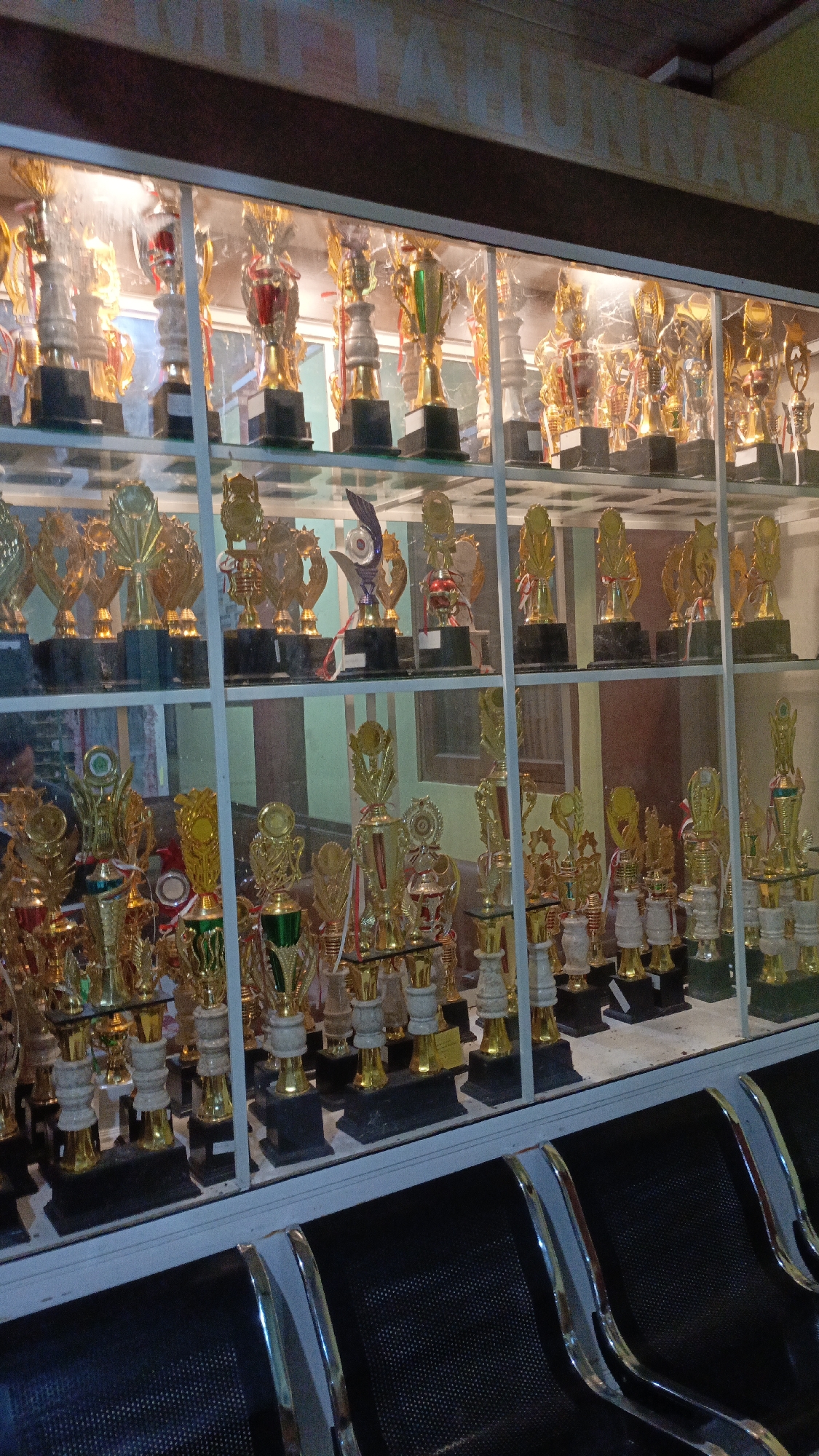 This is a medium-angle, eye-level, indoor shot of a trophy display case filled with numerous gold-colored trophies. The trophies vary in size and design, with some featuring figures, while others are more abstract. The display case is illuminated from within, highlighting the polished surfaces of the trophies. Behind the display case, a light green wall and a brown wooden structure are visible. The bottom of the image shows the backs of several black chairs with chrome accents. The scene appears to be in a public or institutional setting, possibly a school or community center, in Trihanggo, Indonesia.