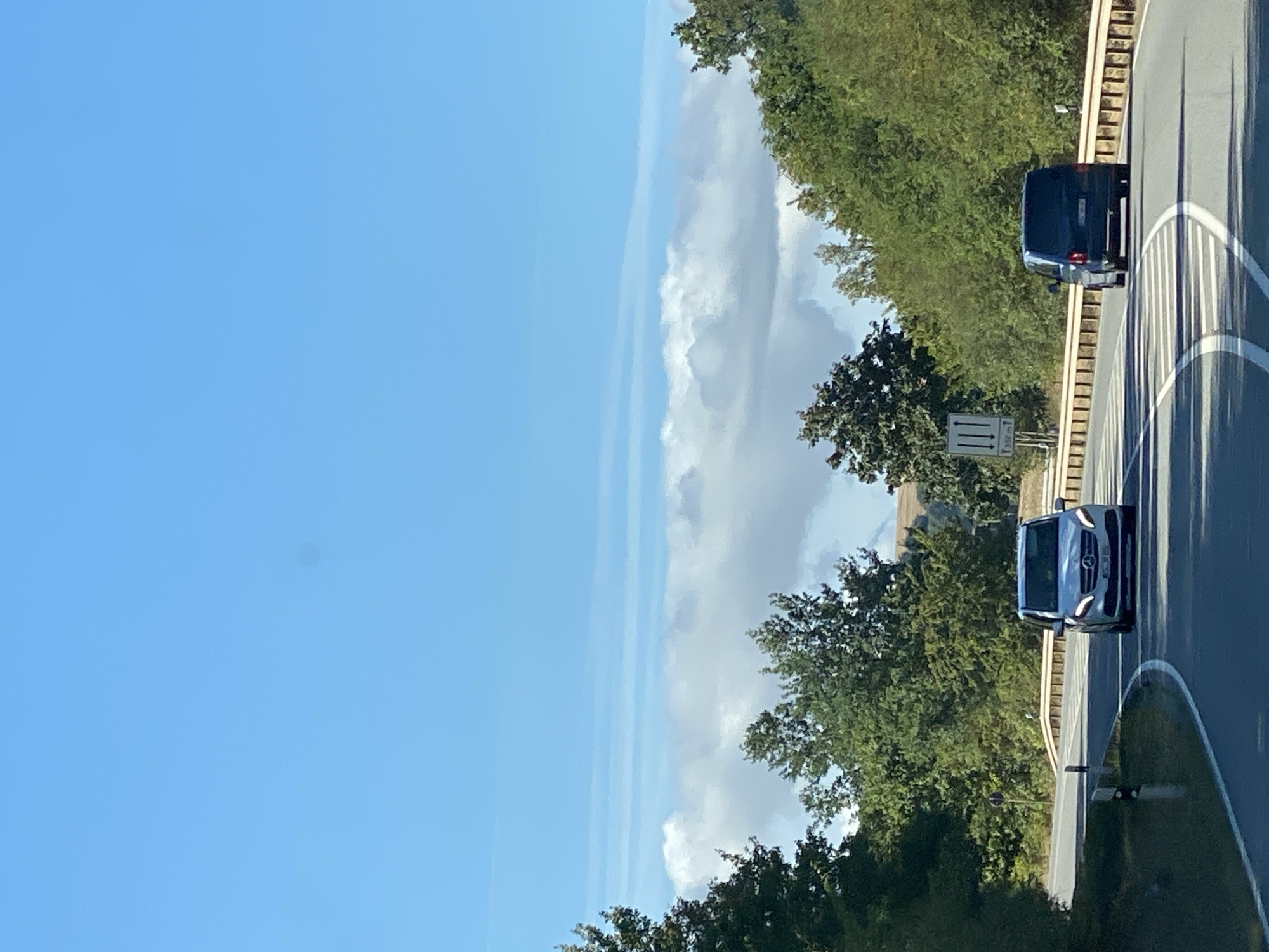 The image depicts a sunny day with a clear blue sky and scattered white clouds over a landscape of green trees. A curved asphalt road is visible, with a light-colored van driving on the right lane and a dark blue van in the left lane, both heading away from the viewer. A yellow guardrail runs along the side of the road, and a road sign indicating 1350m is present on the left. The scene suggests a rural or suburban area, possibly on the outskirts of Nordhausen, Germany, given the presence of a road sign with distance markers, typical of German infrastructure. The overall impression is of a calm, scenic drive.