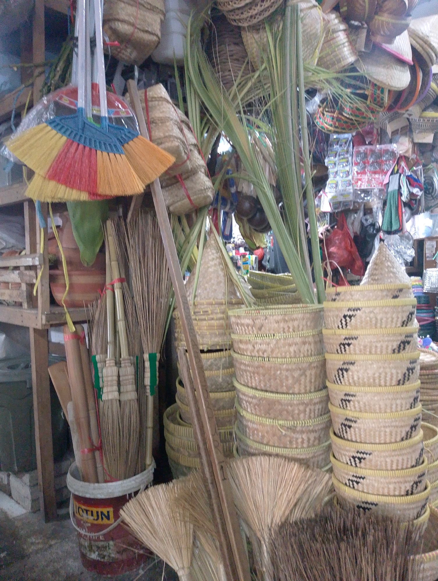 The image depicts a cluttered stall in a marketplace, likely in Lembang, Indonesia, selling a variety of handmade items. The foreground is dominated by brooms and other cleaning tools. Several bundles of brooms made from natural fibers are arranged in a red bucket, with a partially visible logo that reads "CTUN". A wide, fan-shaped broom made of lighter-colored straw lies on the ground in front of the bucket.
Behind the brooms, stacks of woven baskets of various sizes and shapes are prominent. These baskets appear to be made from natural materials like rattan or bamboo, with some featuring decorative black patterns. In the background, more baskets and woven items are visible, along with some plastic-wrapped goods and what appear to be palm leaves. A wooden crate is visible on the left side of the frame, and hanging items, possibly clothing or bags, are present in the upper background. The overall impression is one of a busy, traditional market stall. There are no people clearly visible in the image. The lighting suggests an indoor or covered area, possibly with natural light filtering in.