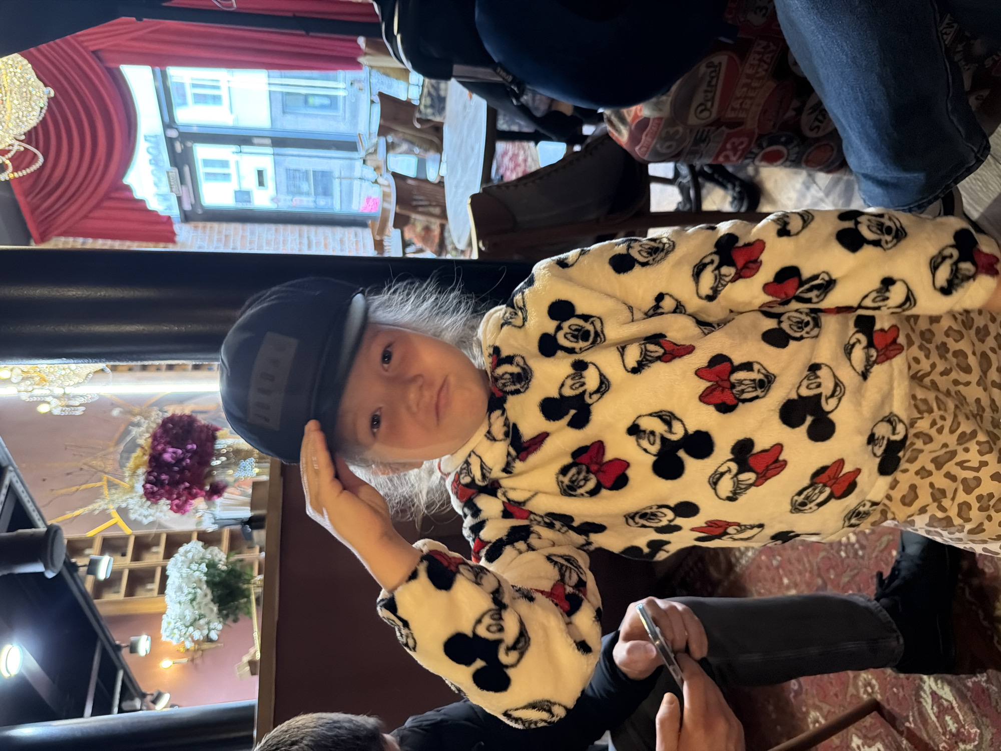 A girl wearing a black baseball cap and a white and black Mickey Mouse print jacket is indoors. The location appears to be a restaurant in Moscow, Russia, based on the window view of buildings and a possible street name in Cyrillic script. The girl is holding the cap's brim. Seated at a table, other people are visible, and a phone is in someone's hand. The restaurant features decorative elements like chandeliers and floral arrangements. The decor includes red curtains and wooden shelves. A mix of furniture and carpeting are also present.