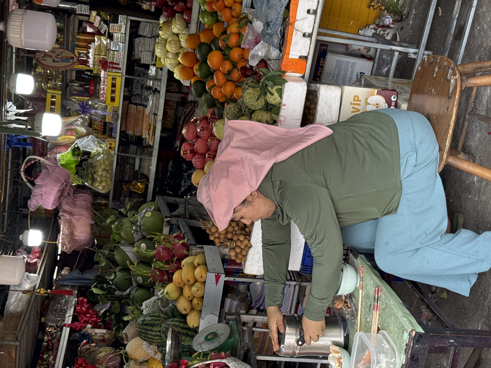 A woman wearing a pink headscarf, light blue trousers, and a long-sleeved green top sits on a wooden stool, operating a metal appliance. She is positioned in front of a table laden with a variety of fruits and vegetables including oranges, dragon fruit, mangoes, watermelons, and other unidentified produce. The background reveals a busy market setting with shelves stocked with diverse goods, suggesting a commercial environment. The location is plausibly a bustling market in Đà Nẵng, Vietnam, given the types of produce and market ambiance depicted.
