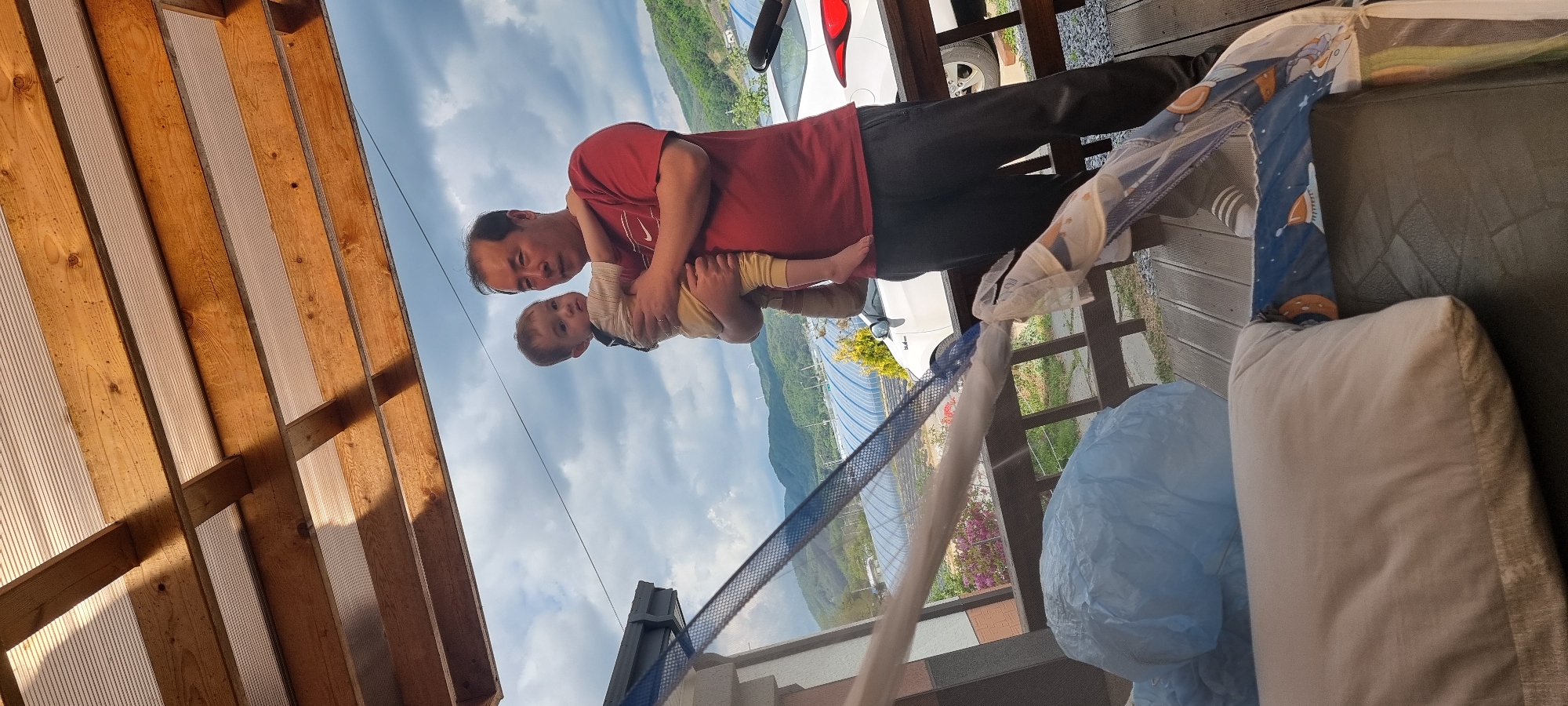 A man holds a young child in his arms outdoors, seemingly on a porch. The man wears a red shirt and dark pants. The child is wearing a light-colored outfit. In the background is a landscape with mountains and a cloudy sky. A white car is partially visible. Elements such as wooden beams, a white cushion, and a blue plastic bag are also visible in the scene. The location is Dunnae-myeon, South Korea.