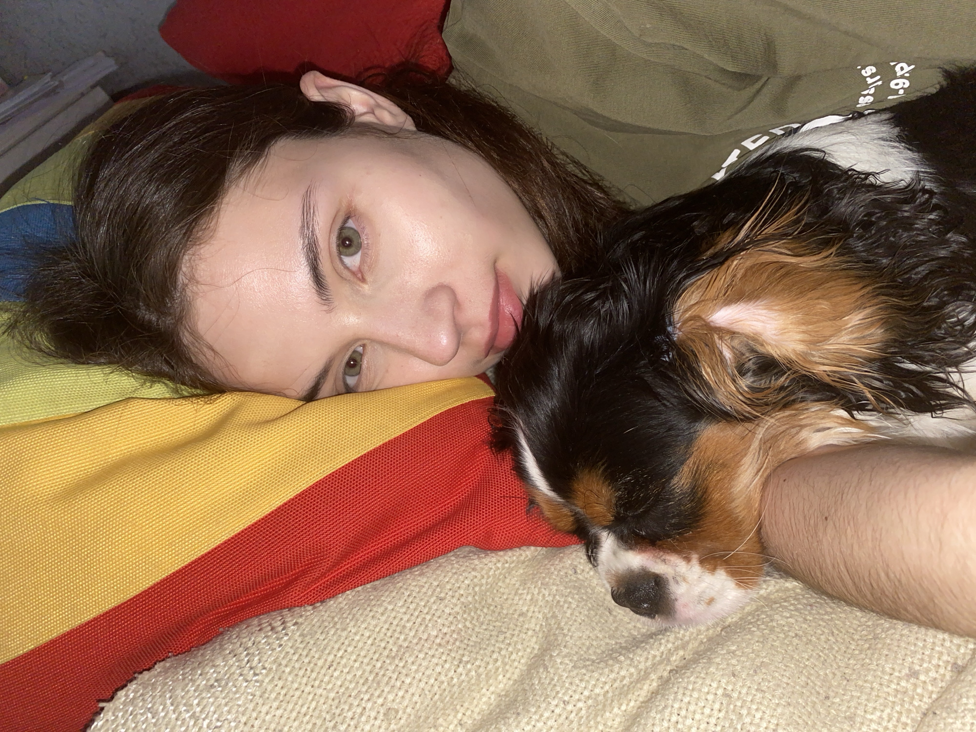An indoor, eye-level close-up shot captures a young person and a small dog resting together in a cozy setting. The person, with dark brown hair and light green eyes, lies on their side, looking towards the camera with a gentle, soft expression. Their head rests on a striped pillow featuring sections of yellow, red, and blue/denim fabric.
Nestled closely against the person's arm and shoulder, a tri-color dog, likely a Cavalier King Charles Spaniel with black, tan, and white fur, is asleep. The dog's eyes are closed, and its head is comfortably tucked, conveying a sense of deep peace. Both subjects are partially covered by a cream-colored, chunky knit blanket or throw, which adds to the comfortable atmosphere. A red pillow is visible behind the person's head, and they are wearing an olive green top.
The dimly lit environment suggests a relaxed moment, possibly during the evening or night. The overall mood is calm, peaceful, and affectionate, highlighting the bond between the person and the dog. Partially visible white text, which appears to read "SIR`S 1-9.D" or similar, is present on the olive green fabric behind the dog. The location context provided is Votkinsk, Russia.