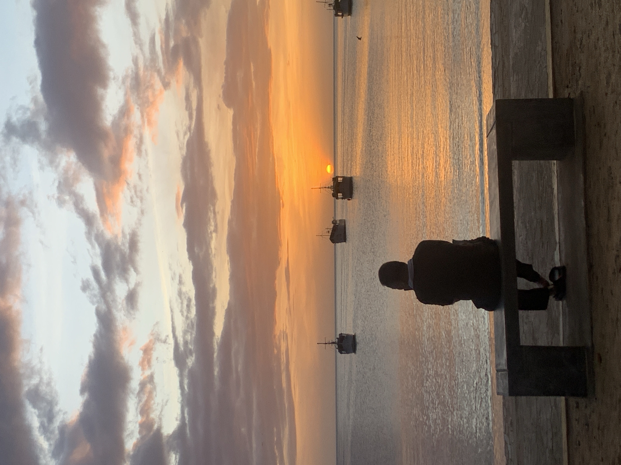 A lone figure is seated on a dark, concrete bench, facing away from the camera, towards a body of water during sunset. The individual is wearing dark clothing and appears to be looking out at the horizon. Several boats are visible on the water, some of which are positioned closer to the shore, while others are further out. The sky is filled with colorful clouds, ranging from shades of orange and pink to gray and blue, indicating a vibrant sunset. The water reflects the colors of the sky, creating a shimmering effect. The foreground of the image shows the bench and a sandy or paved area. The overall scene suggests a peaceful and contemplative moment at the shore of Juan Griego, Venezuela.