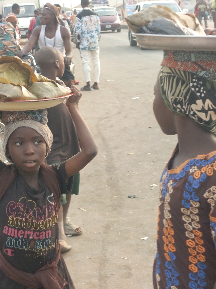The image depicts a street scene in Alizaga, Nigeria, characterized by pedestrian and vehicular activity. In the foreground, a young dark-complexioned girl is positioned left of center, looking slightly upward and to her left. She wears a dark t-shirt with multicolored "Champion authentic american athletic" text, a brown wrap tied around her waist, and a light-colored head covering. She balances a red-rimmed white tray on her head, laden with multiple items wrapped in leaves or paper, supporting it with her raised right hand. To her right, the upper torso and head of another individual are visible, wearing a brown garment with blue and orange embroidered patterns and a multi-patterned headscarf. This person also carries a metal tray on their head, holding what appears to be a large piece of dried fish or meat and other packaged items.
In the midground, several other individuals are present. A dark-complexioned man in a white tank top and a baseball cap stands near a bicycle, looking left. Further back, another man is seen in a white shirt with a tropical pattern and white trousers. The background contains additional blurry figures and several vehicles, including a white van and two cars on what appears to be a paved road. The ground surface is a worn, light-colored pavement with sparse debris. The scene is illuminated by natural daylight.
