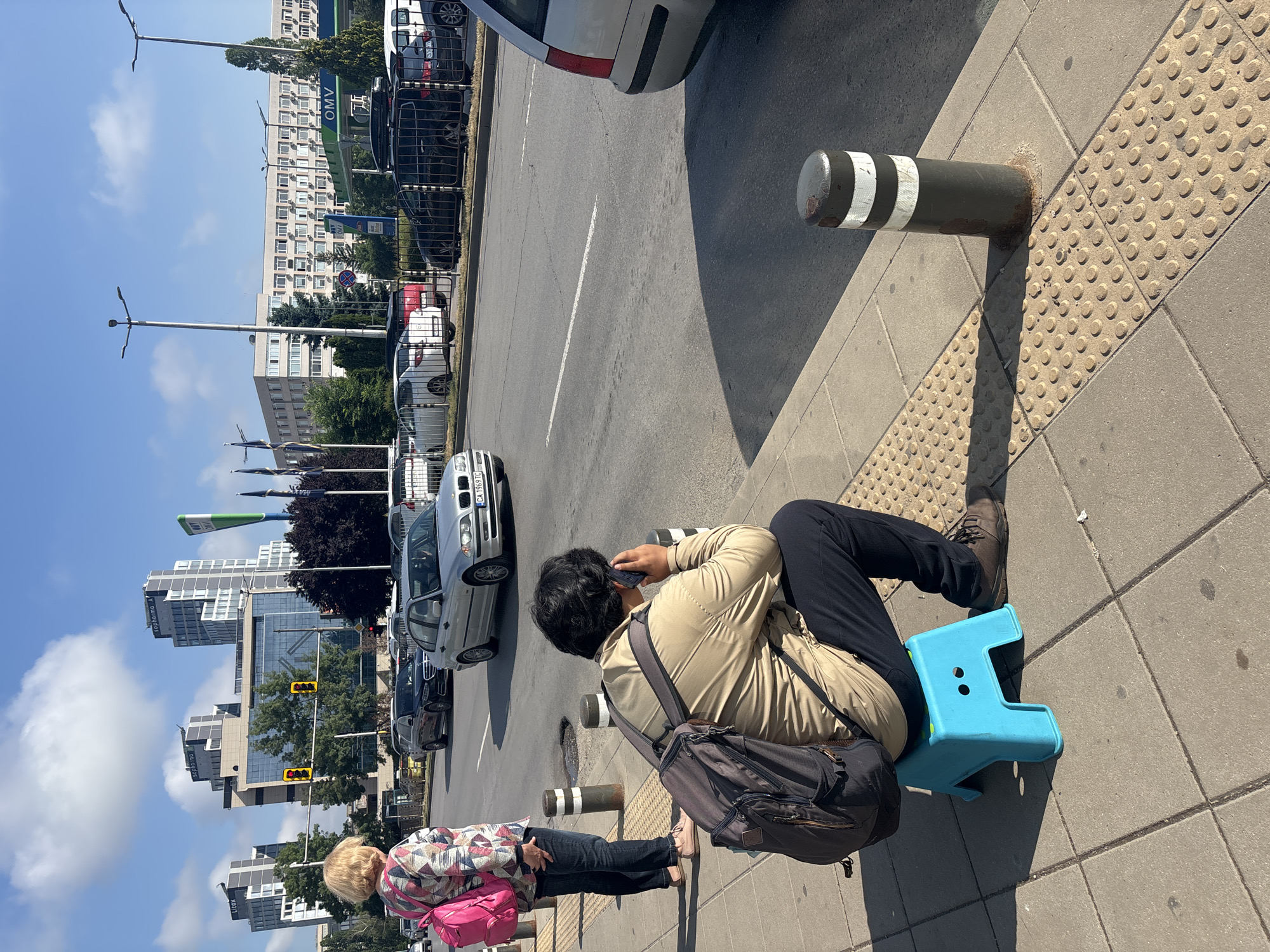 The image depicts a vibrant urban street scene under bright, sunny conditions. In the foreground, a person with dark hair, wearing a beige jacket and black pants, is seated on a bright blue plastic stool on the sidewalk. They are holding a dark smartphone to their ear, appearing to be on a call or listening intently. A dark shoulder bag with a brown flap is slung across their body. Next to them, a second individual with blonde hair and a patterned top stands, carrying a bright pink shoulder bag.
The sidewalk features light-colored paving stones and sections of tactile paving with raised dots, commonly used for the visually impaired. Several dark bollards with white reflective stripes are embedded along the curb. Strong, well-defined shadows are cast by the people and objects, indicating bright sunlight, with a clear blue sky and a few white clouds visible above.
Beyond the sidewalk, a multi-lane asphalt road with white markings is visible, where several cars are either parked or in motion. A silver car prominently displays a Bulgarian license plate, "CA 1969 TC". Tall streetlights with multiple lamp heads line the street. In the background, a mix of architectural styles is present, including multi-story residential or office buildings and modern high-rises with glass facades. Visible text on the buildings includes "saphirno" and "CLICK". An "OMV" sign, likely for a gas station, is also discernible amidst the buildings and trees.