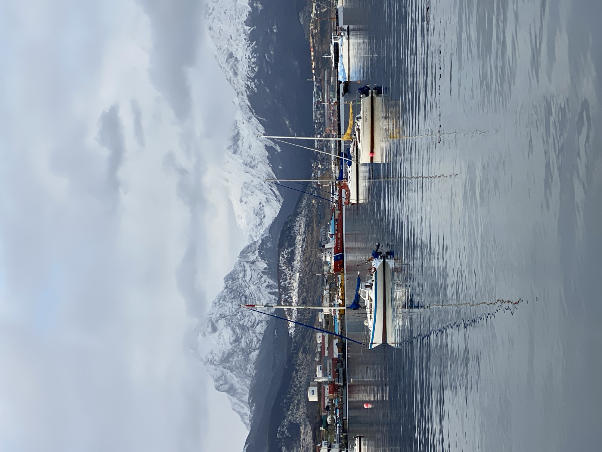 The image depicts a harbor scene in Ushuaia, Argentina, characterized by a gray and overcast sky. In the background, snow-capped mountains are partially obscured by clouds. Several sailboats are moored in the calm water, creating vertical reflections in the water. A red buoy is visible near the boats. The waterfront is lined with buildings, including shipping containers and structures of varying sizes and colors. The overall scene presents a cold and serene atmosphere.
