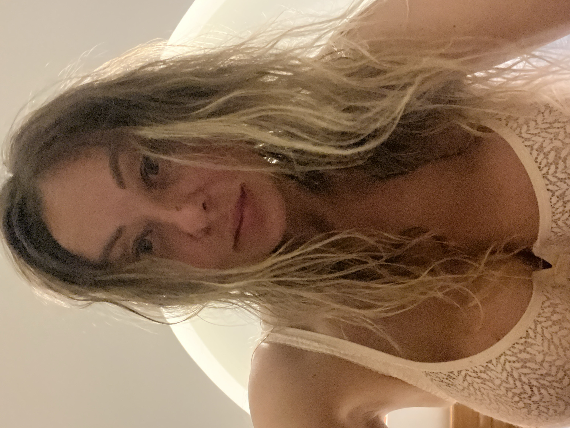 A low-angle, close-up, head-and-shoulders shot of a woman with wet, shoulder-length blonde hair, wearing a cream-colored lace bra. She is looking directly at the camera with a neutral expression. The background is a plain, light-colored wall with a subtle arch shape visible on the left. The lighting is soft and diffused. The location of this image is not specified within the image itself or the provided context.